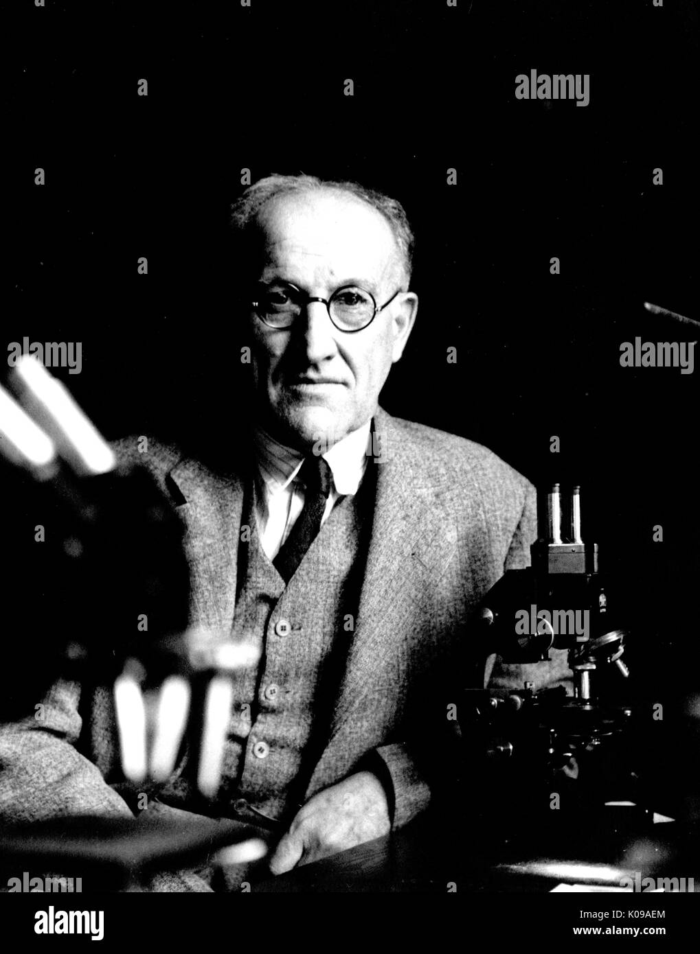 Half-length photograph of Dr. Samuel Mast, biology faculty member at the Johns Hopkins University, sitting behind a wooden desk, wearing a dark three piece suit and circular glasses, a microscope and other instruments on the table in front of him, with a serious facial expression. 1937. Stock Photo