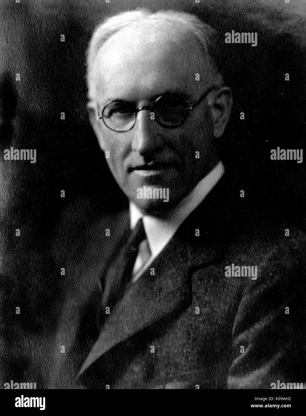 Half-length of Dr. Samuel Mast, biology faculty member at the Johns Hopkins University, wearing a dark three piece suit and a dark tie with a white shirt and circular dark glasses, in front of a backdrop, with a serious facial expression. 1935. Stock Photo