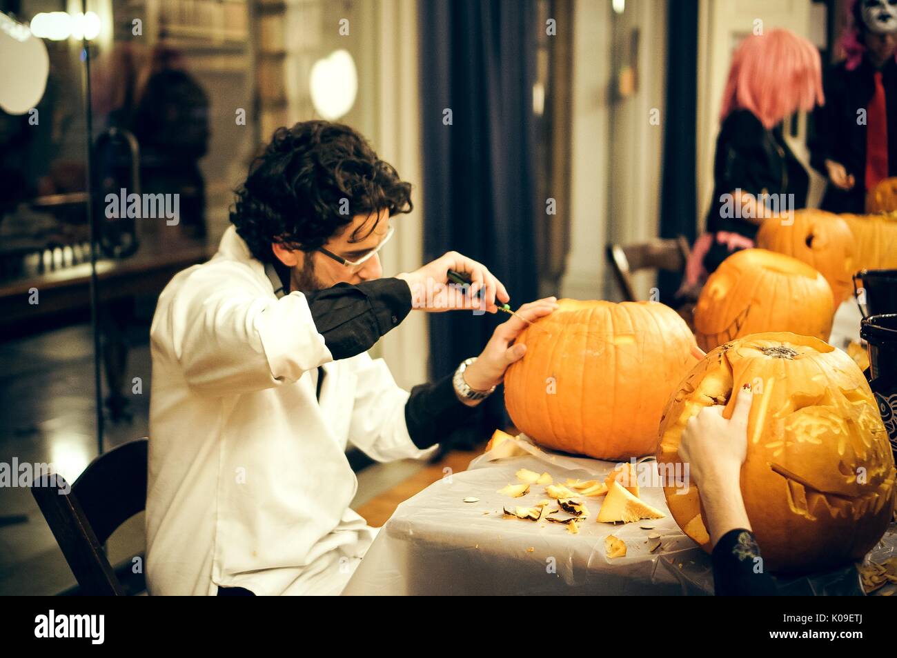 Student seated around table carving a pumpkin with a knife, surrounded by other college students carving faces into pumpkins, student wearing a white smock atop clothing, Peabody Halloween party, October 31, 2015. Courtesy Eric Chen. Stock Photo