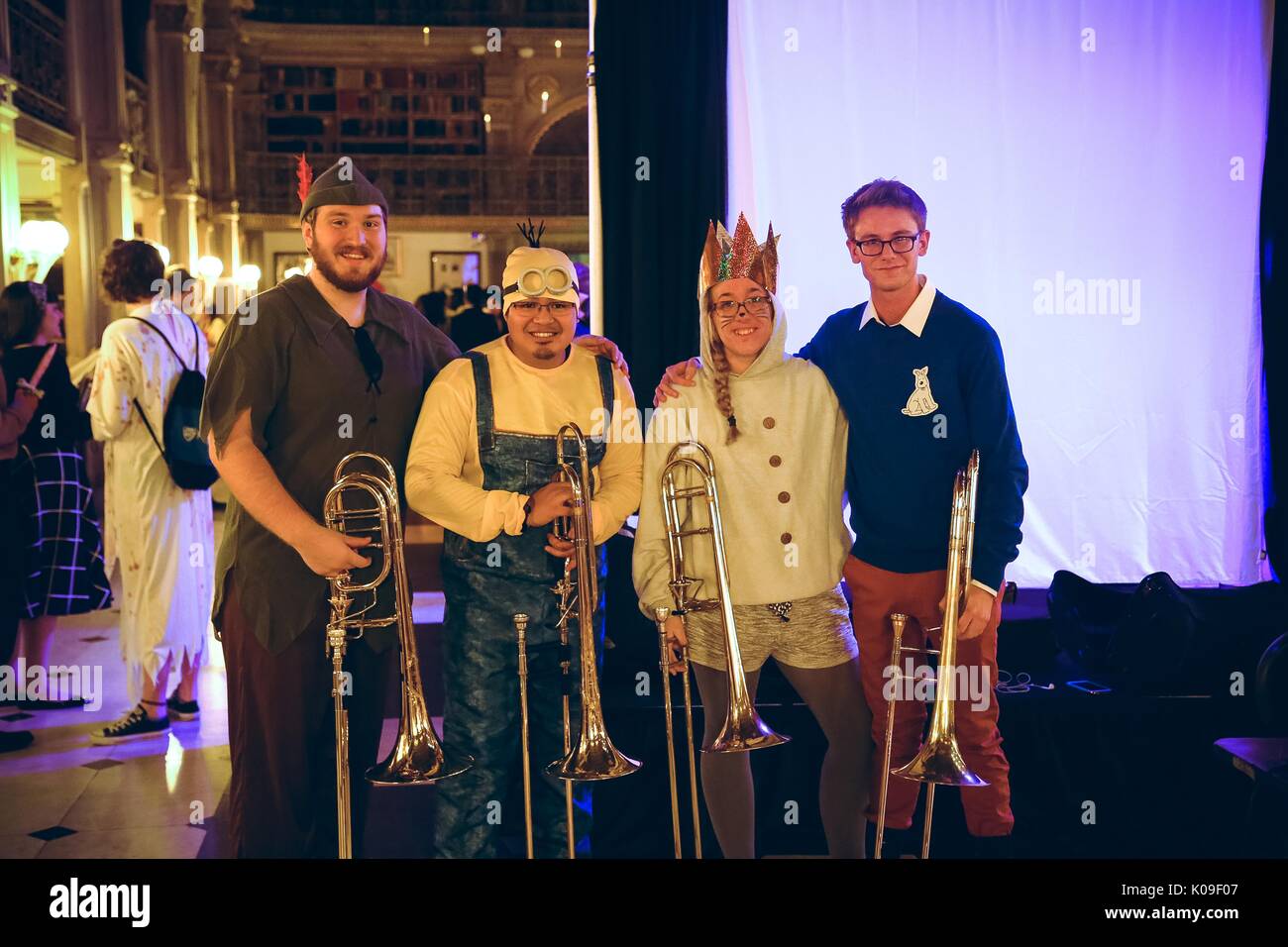 Four students all holding trombones dressed in costumes, the leftmost male student dressed as Peter pan, the male student second from the left dressed as a Minion from the film 'Despicable Me'; the female student second from the rights wearing a shining crown and cat make up, the rightmost male student wearing a blue sweater with a drawing pinned onto his chest and orange pants; Halloween at Peabody, October 31, 2015. Stock Photo