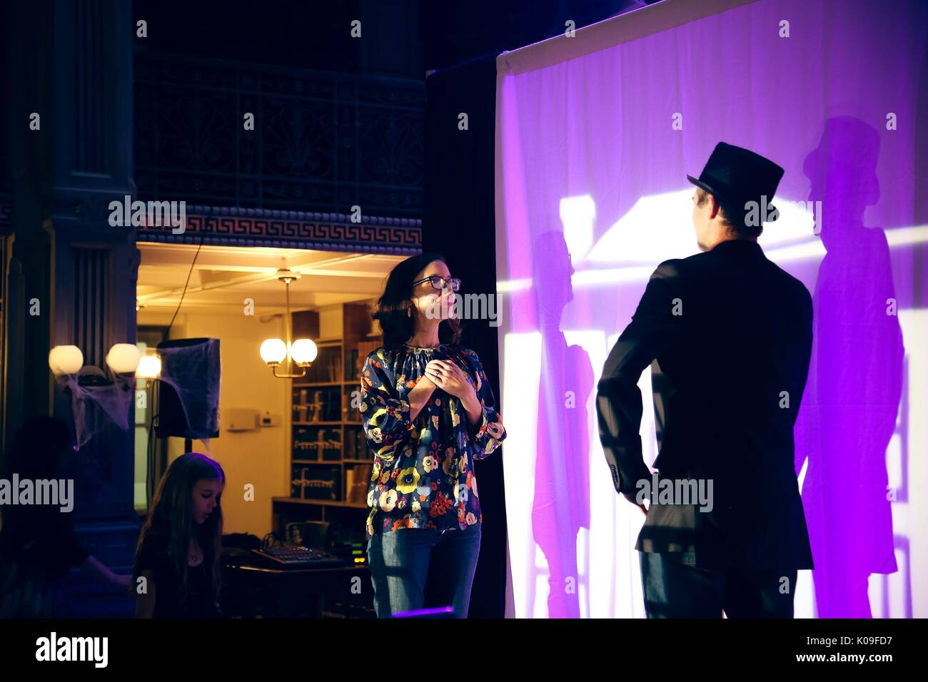 A man in a suit and top hat and a woman in regular clothing are on a stage, spot lights are on them and they are in front of a white cloth screen that captures their shadows, they are looking towards each other, 2015. Courtesy Eric Chen. Stock Photo