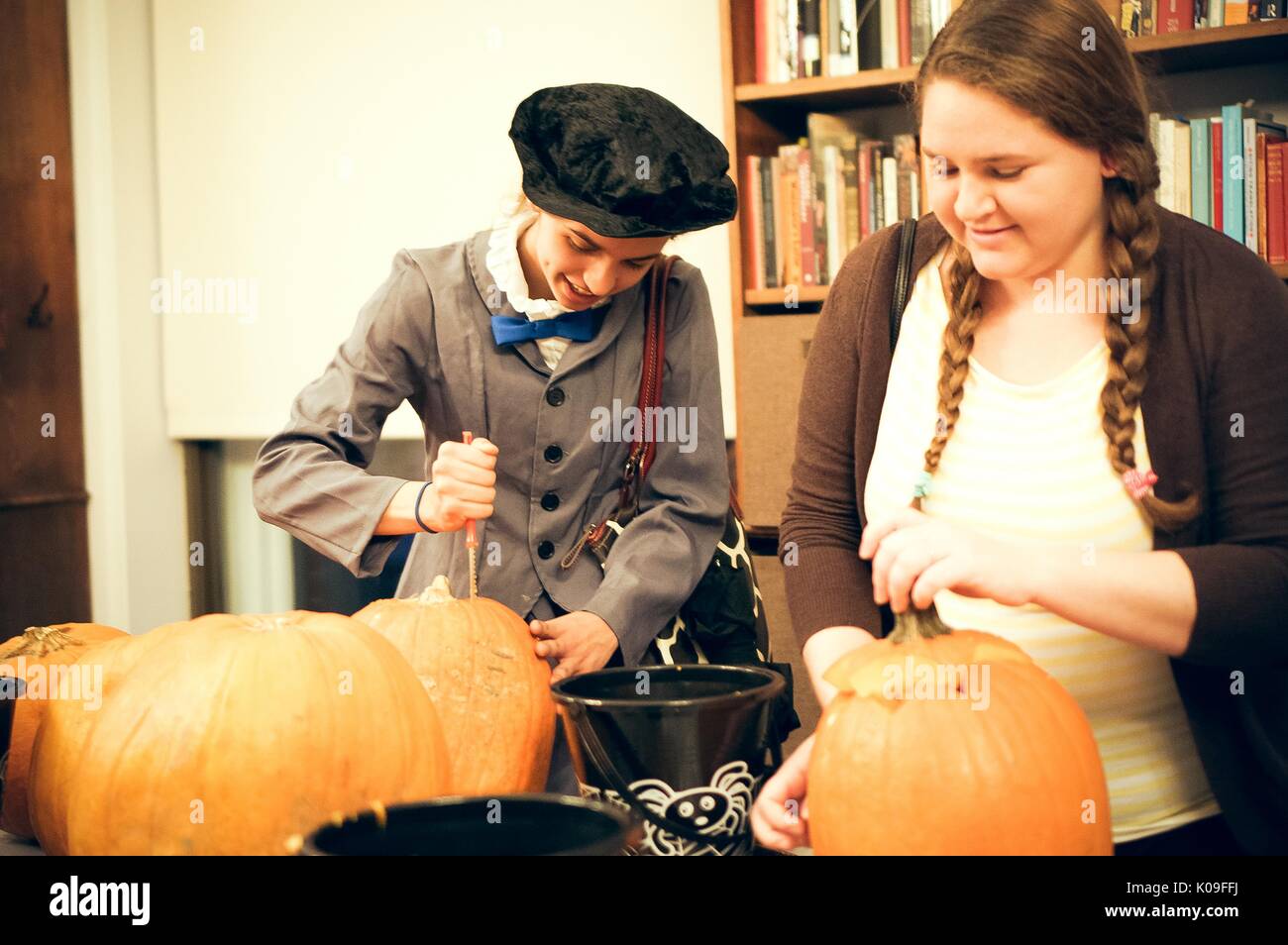 Two college students are carving pumpkins, there are several other pumpkins on the table and several black buckets to collect the scraps, one of the students is in costume wearing a buttoned up suit jacket and bow tie and a dark-colored hat, 2015. Stock Photo