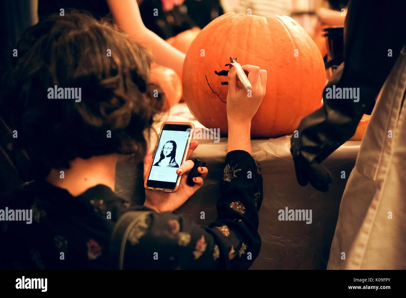 A college student is concentrating on drawing the Mona Lisa on her pumpkin as she holds an image of it on her phone in front of her, 2015. Courtesy Eric Chen. Stock Photo
