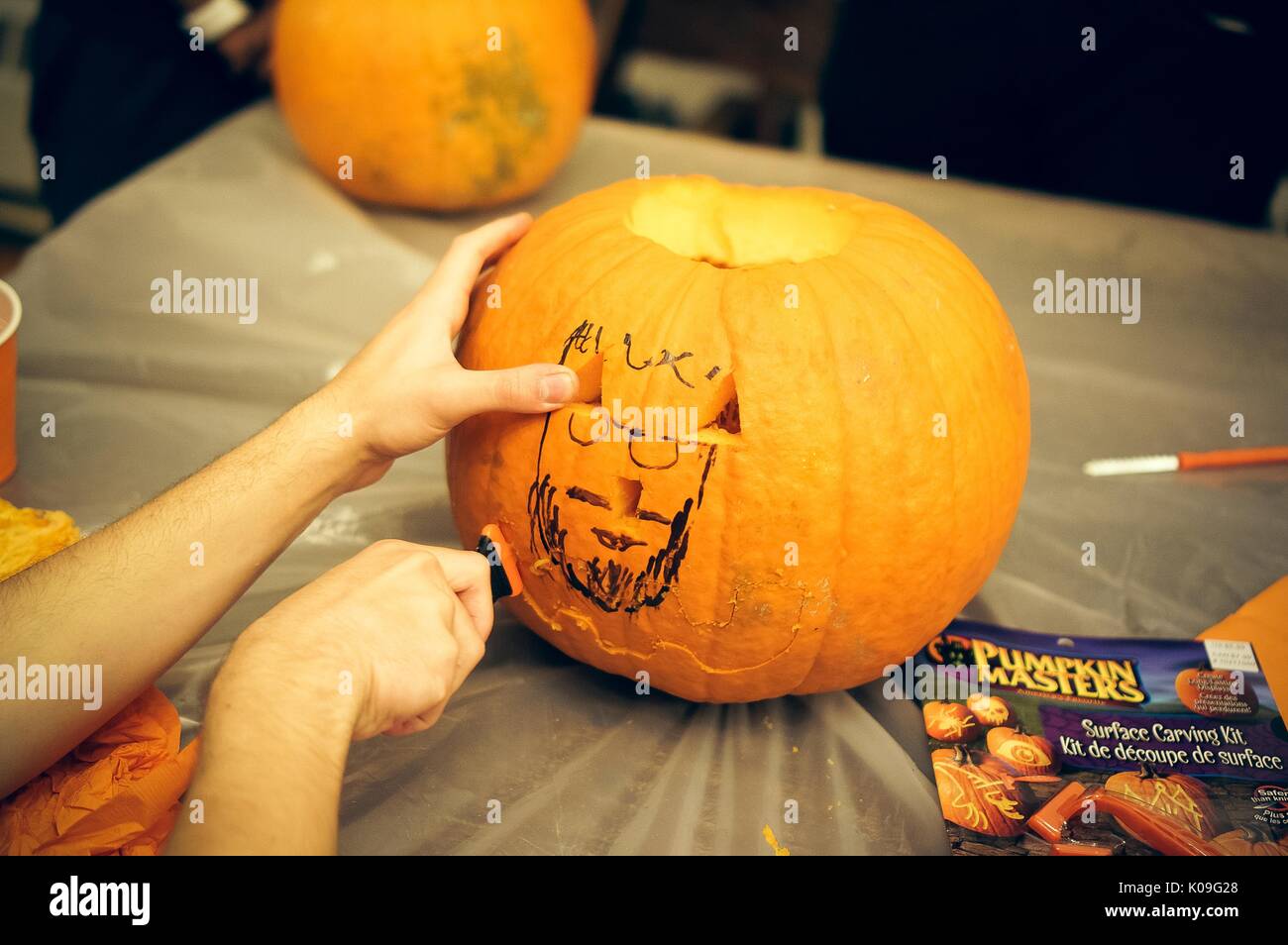 View of the front of a pumpkin that has had its stem and some of its side carved out, one hand is holding the pumpkin while the other works to carve the pumpkin, there is a drawing of a face on the pumpkin with Sharpie, 2015. Stock Photo