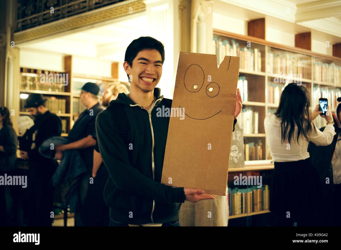 A college student posing for a picture, he is smiling and holding a board that has two holes in it for eyes and a drawn-on smile, Halloween at Johns Hopkins University's George Peabody Library, 2015. Courtesy Eric Chen. Stock Photo