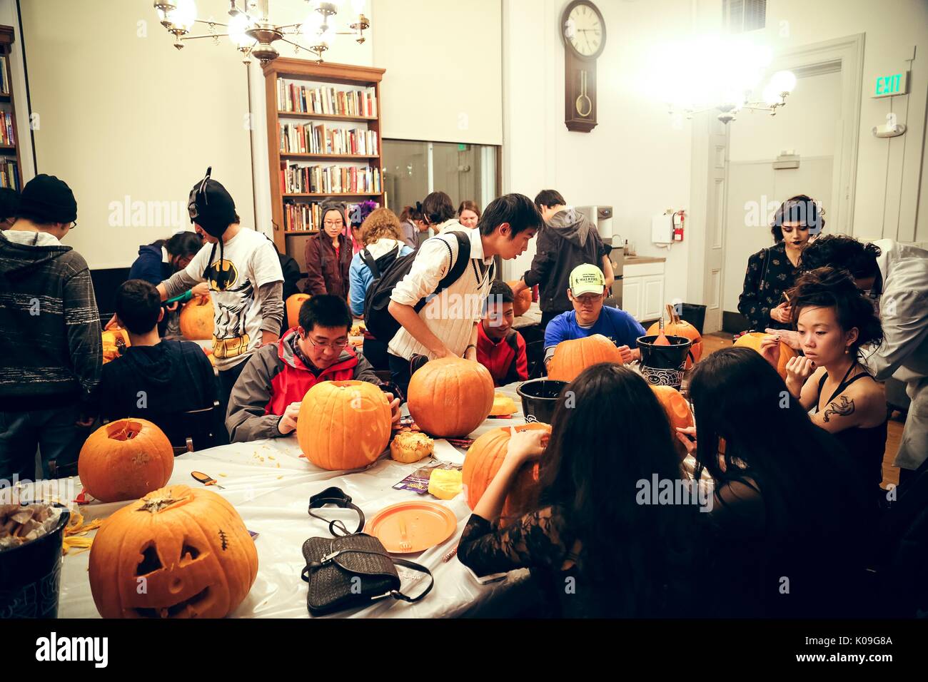 College students are in a room that is dedicated to carving pumpkins, they are all dressed in different costumes and are sitting and standing working on their pumpkins at two long rectangular tables covered in pumpkins, Halloween at Johns Hopkins University's George Peabody Library, 2015. Courtesy Eric Chen. Stock Photo