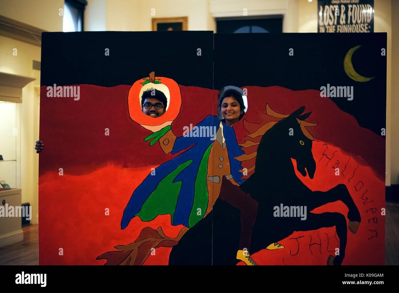 Two college students pose behind a face-in-a-hole board, the board has a horse and a rider on it and the rider is holding a pumpkin, the two holes for faces are in the pumpkin and the head of the rider, the board says 'Halloween JHU' on it, 2015. Courtesy Eric Chen. Stock Photo