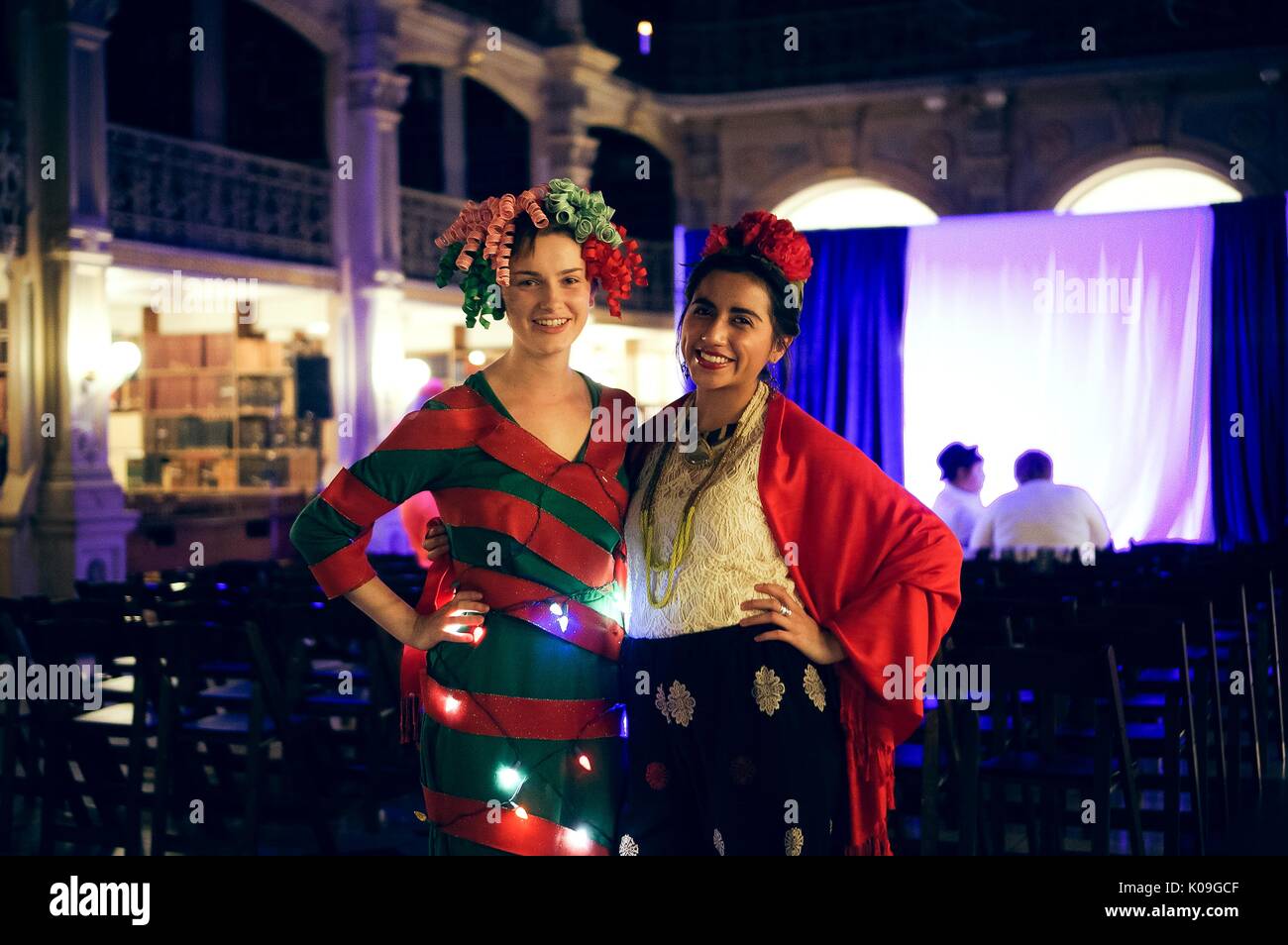 Two female college students pose for a picture, both are in costume, one is wearing a red and green striped outfit and a wig made of ribbons and the other is wearing a red shawl and a red prop in her hair, Halloween at Johns Hopkins University's George Peabody Library, 2015. Courtesy Eric Chen. Stock Photo