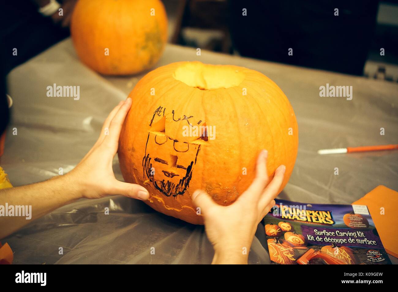 View of the front of a pumpkin that has had its stem and some of its side carved out, there are two hands holding the pumpkin on an angle and a drawing of a face is on the pumpkin with Sharpie, 2015. Courtesy Eric Chen. Stock Photo