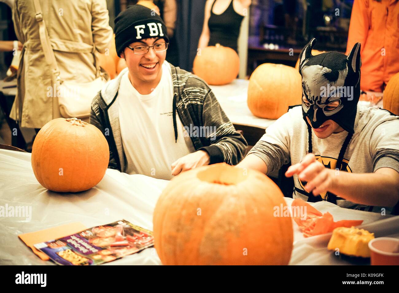 Two college students are sitting at a table each with a pumpkin, one is carving their pumpkin and the other pumpkin has not been touched yet, the students are smiling and are also in costume, one is wearing an FBI hat and the other is wearing a Batman hat and mask, Halloween at Johns Hopkins University's George Peabody Library, 2015. Courtesy Eric Chen. Stock Photo