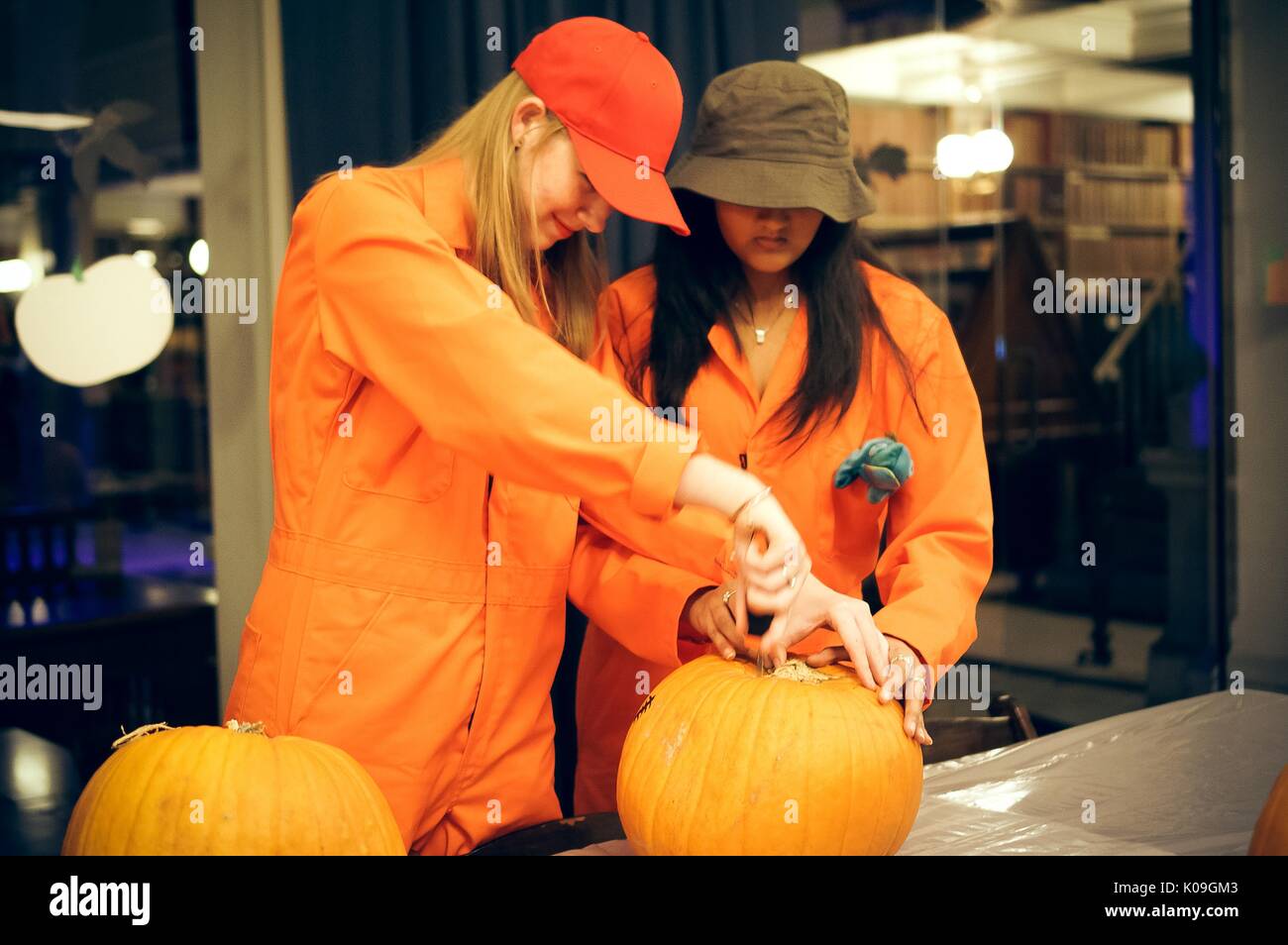 Two college students work together at carving the stem out of one pumpkin, they are dressed in orange jumpsuits as characters from the story 'Holes', Halloween at Johns Hopkins University's George Peabody Library, 2015. Courtesy Eric Chen. Stock Photo