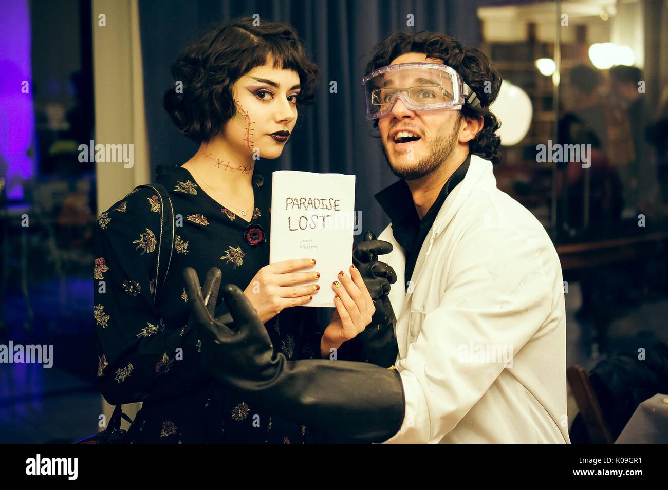 Two college students pose for a picture, the girl has fake drawn-on stitches on her face and is holding a prop with the title 'Paradise Lost' and the boy is dressed as a scientist and is wearing a lab coat, goggles, and gloves, 2015. Courtesy Eric Chen. Stock Photo