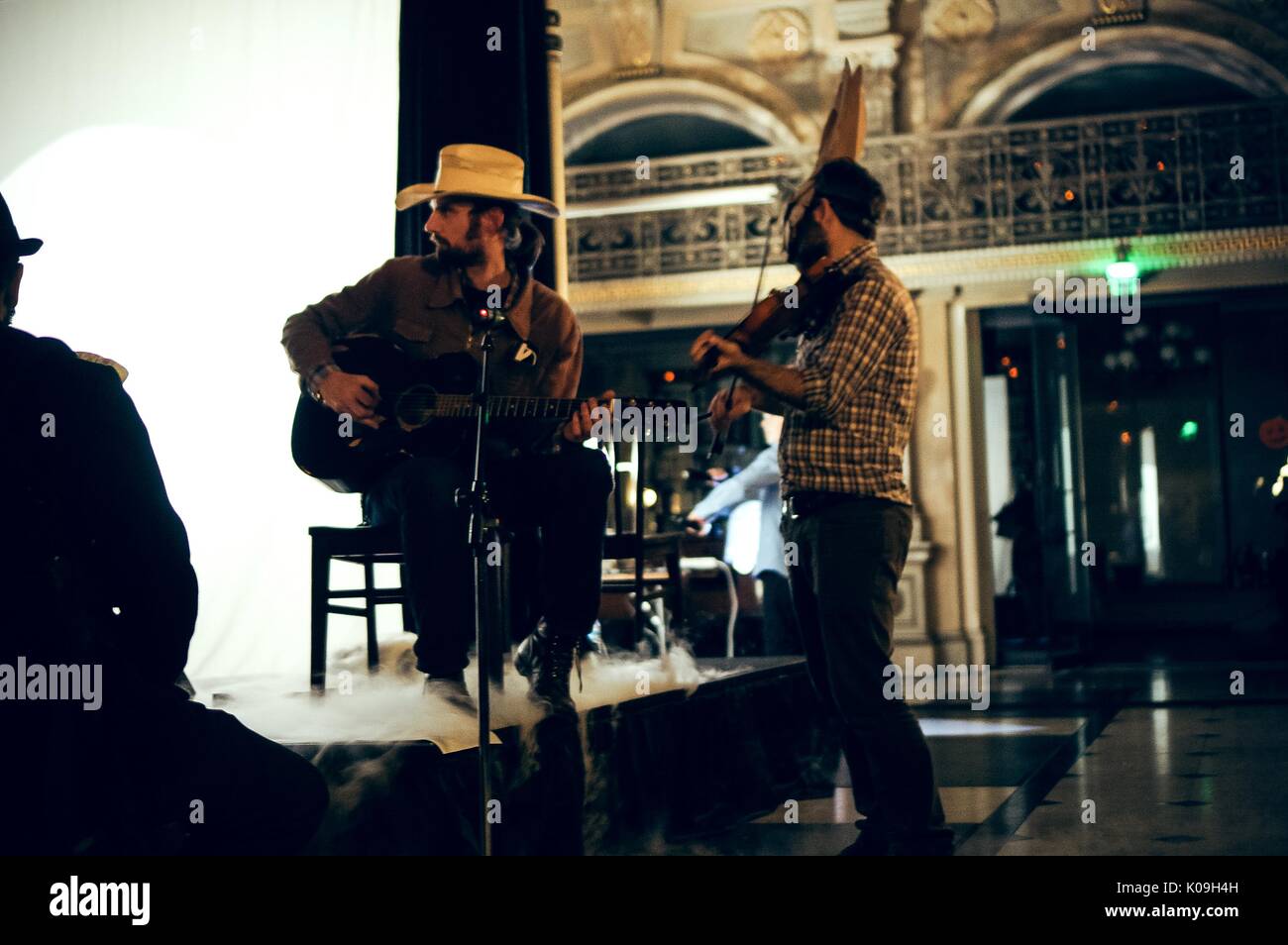 Two musicians are by a stage, on Halloween at Johns Hopkins University's George Peabody Library, one man is sitting on a chair on the stage with a guitar in his lap and the other man is holding a violin, 2015. Courtesy Eric Chen. Stock Photo