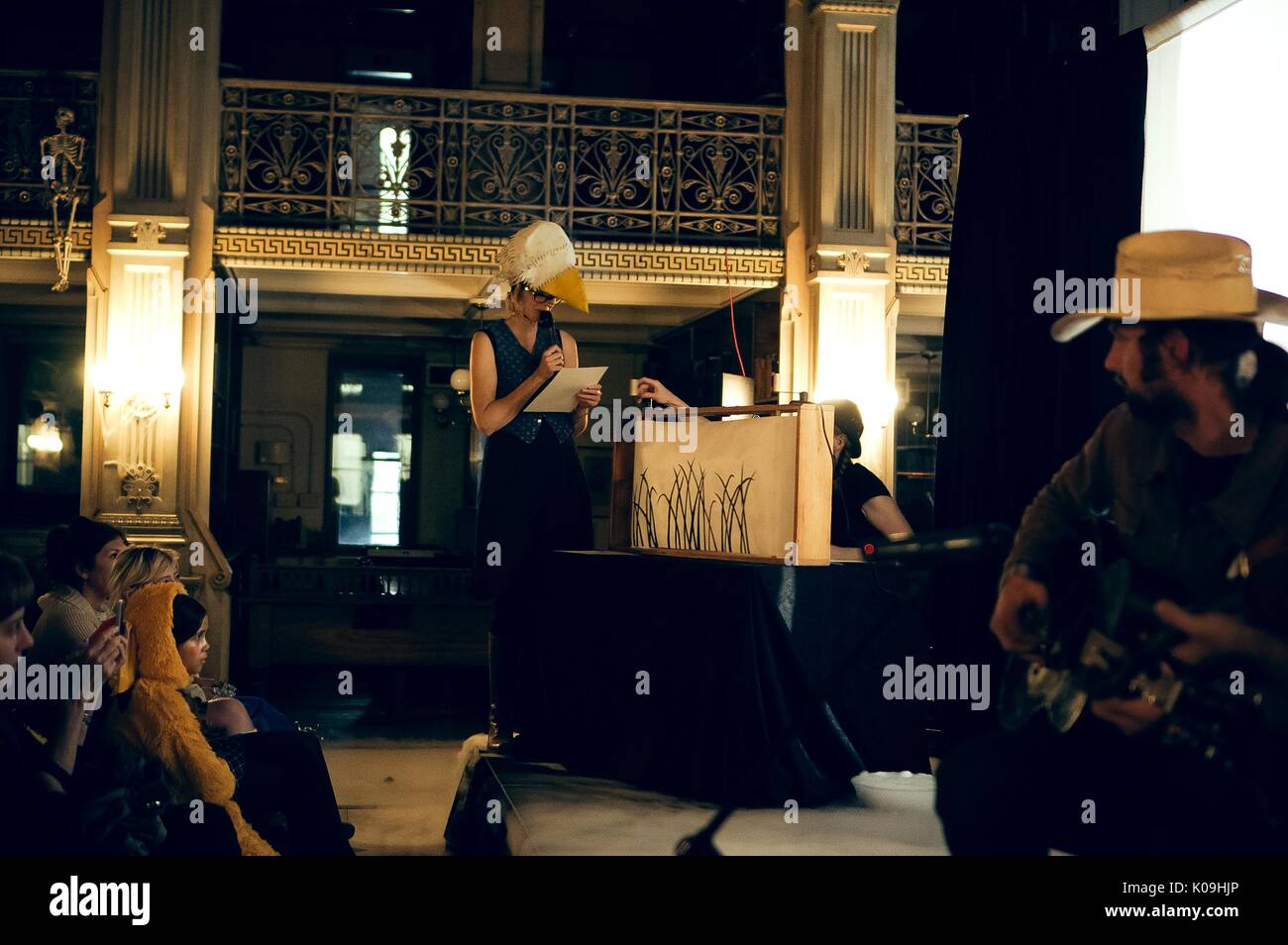 Side angle view of a shadow puppet show with narrator wearing a crow-like head piece prop, a musician is in the foreground, 2015. Courtesy Eric Chen. Stock Photo