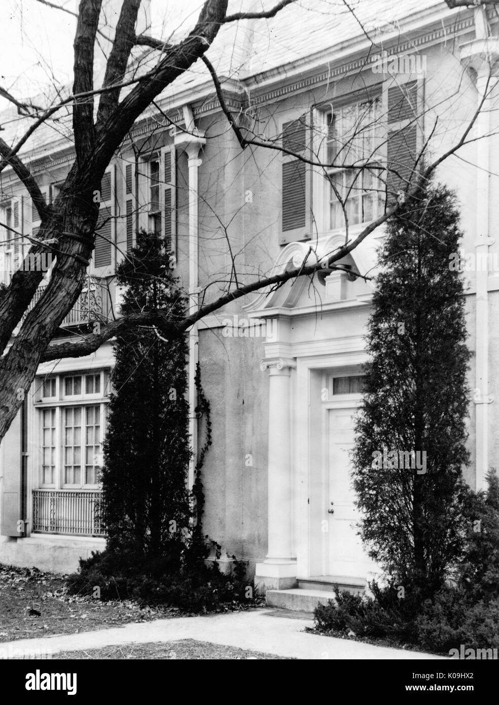 Picture of a facade of a Guilford home in Baltimore, with an ornate front door in between two large shrubs, a two-story home with large windows, Baltimore, Maryland, 1920. This image is from a series documenting the construction and sale of homes in the Roland Park/Guilford neighborhood of Baltimore, a streetcar suburb and one of the first planned communities in the United States. The neighborhood was segregated, and is considered an early example of the enforcement of racial segregation through the use of restricted covenants. Stock Photo