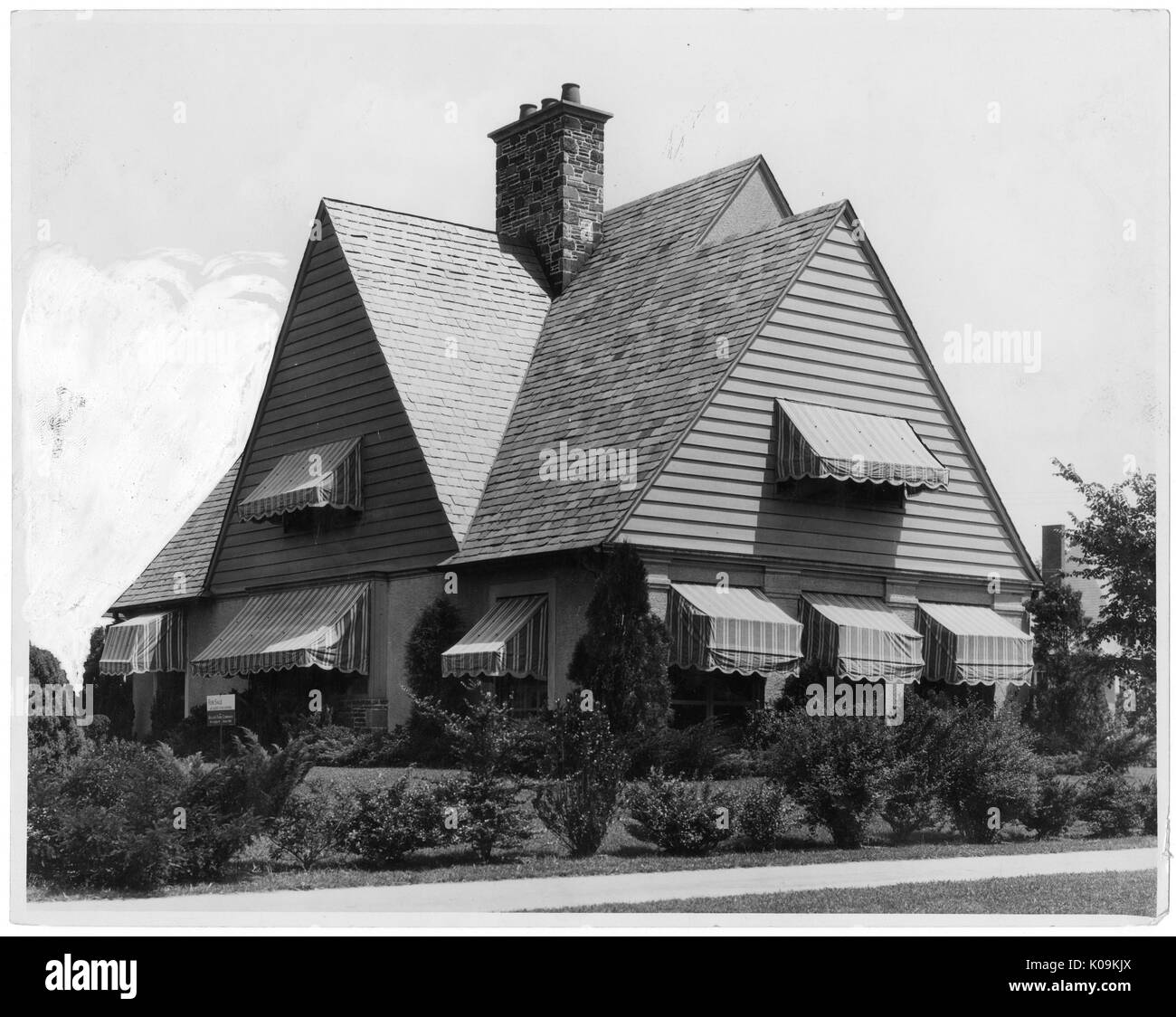 Landscape shot of a stylized two-story house with large, steep triangular roof structures, with window hoods of striped pattern on every window, surrounded by shrubbery arranged in a line, Roland Park/Guilford, Baltimore, Maryland, 1910. This image is from a series documenting the construction and sale of homes in the Roland Park/Guilford neighborhood of Baltimore, a streetcar suburb and one of the first planned communities in the United States. The neighborhood was segregated, and is considered an early example of the enforcement of racial segregation through the use of restricted covenants. Stock Photo