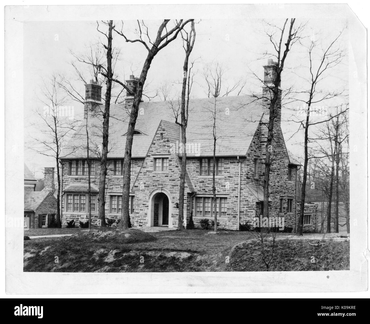 Landscape shot of a large, stone house with two chimneys, a stylized entrance, no landscaping surrounding the building immediately, tall trees without leaves nearby, Roland Park/Guilford, Baltimore, Maryland, 1910. This image is from a series documenting the construction and sale of homes in the Roland Park/Guilford neighborhood of Baltimore, a streetcar suburb and one of the first planned communities in the United States. The neighborhood was segregated, and is considered an early example of the enforcement of racial segregation through the use of restricted covenants. Stock Photo