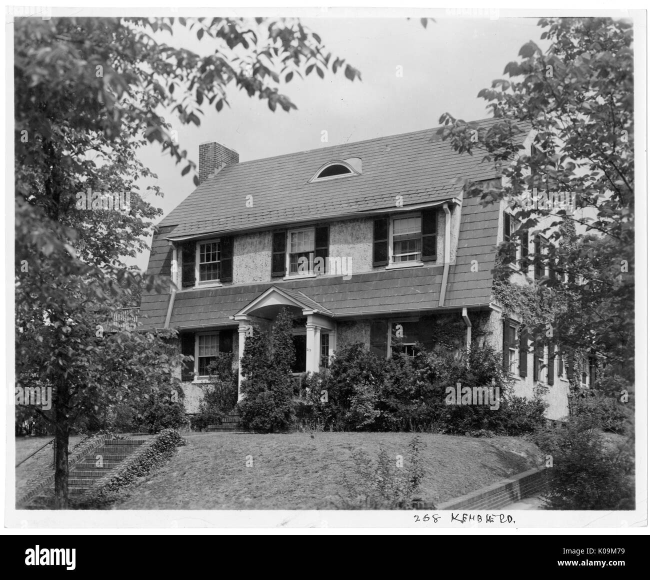 Landscape shot of a two-story house with steps leading to the front door, with a brick chimney, surrounded by shrubs and trees with leaves, Roland Park/Guilford, Baltimore, Maryland, 1910. This image is from a series documenting the construction and sale of homes in the Roland Park/Guilford neighborhood of Baltimore, a streetcar suburb and one of the first planned communities in the United States. The neighborhood was segregated, and is considered an early example of the enforcement of racial segregation through the use of restricted covenants. Stock Photo