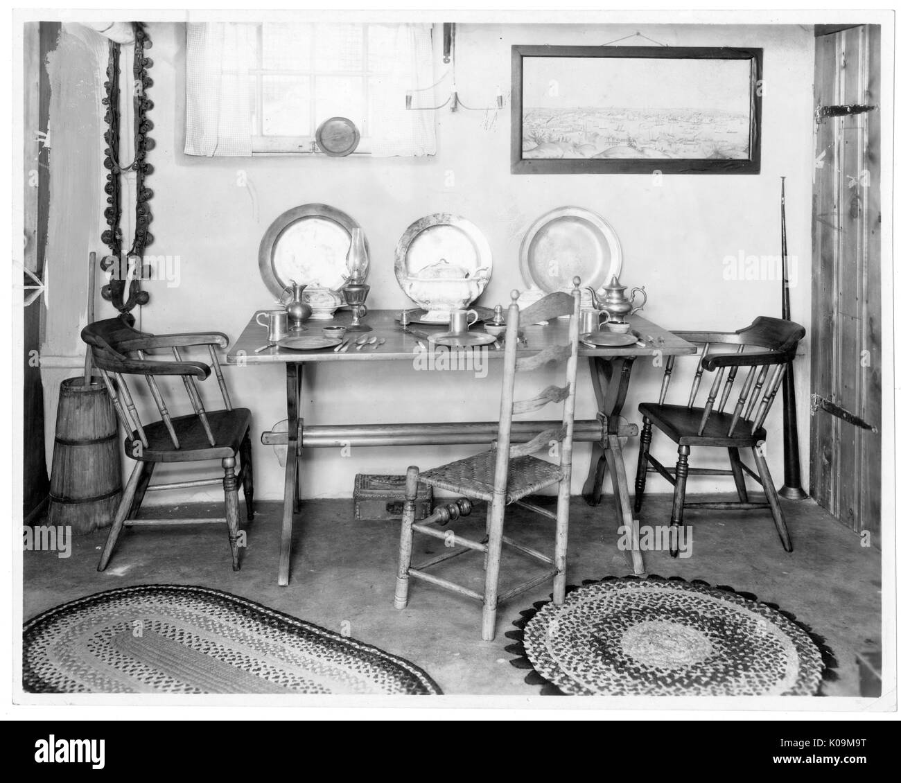 Landscape shot of a wooden table with three different types of wooden chairs; three plate a cup sets lay on the table and propped up against the wall; two different-seized patterned rugs on the ground, a landscape photo hanging on the ceiling next to a large window; Roland Park/Guilford, Baltimore, Maryland, 1910. This image is from a series documenting the construction and sale of homes in the Roland Park/Guilford neighborhood of Baltimore, a streetcar suburb and one of the first planned communities in the United States. The neighborhood was segregated, and is considered an early example of t Stock Photo