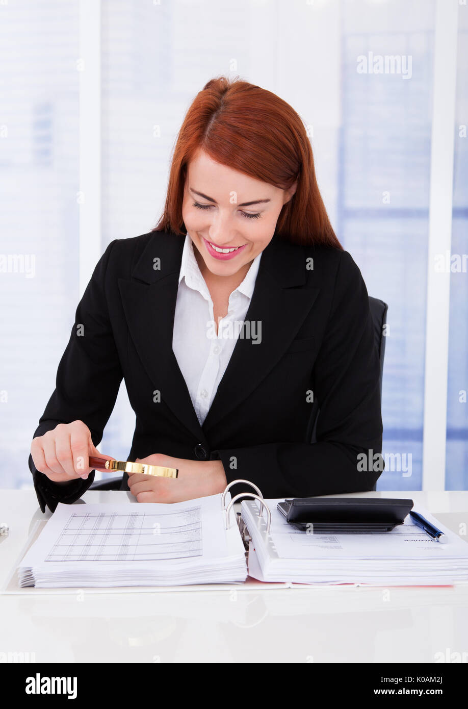Smiling young businesswoman examining documents with magnifying glass at office desk Stock Photo