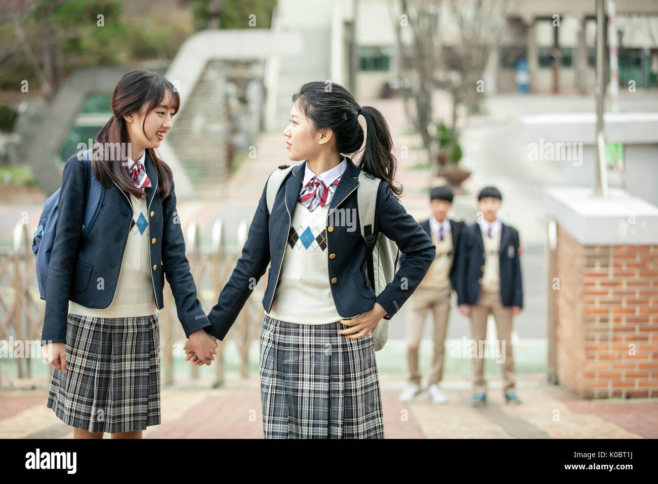 Two smiling school girls standing holding hands face to face against their school Stock Photo