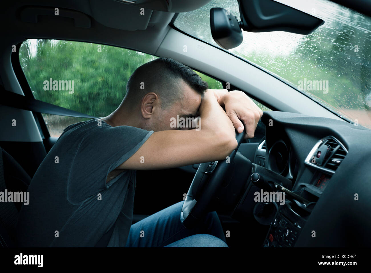 closeup of a young caucasian man sitting at the driver seat of a car relaxing with his head on the steering wheel, in a rainy day Stock Photo