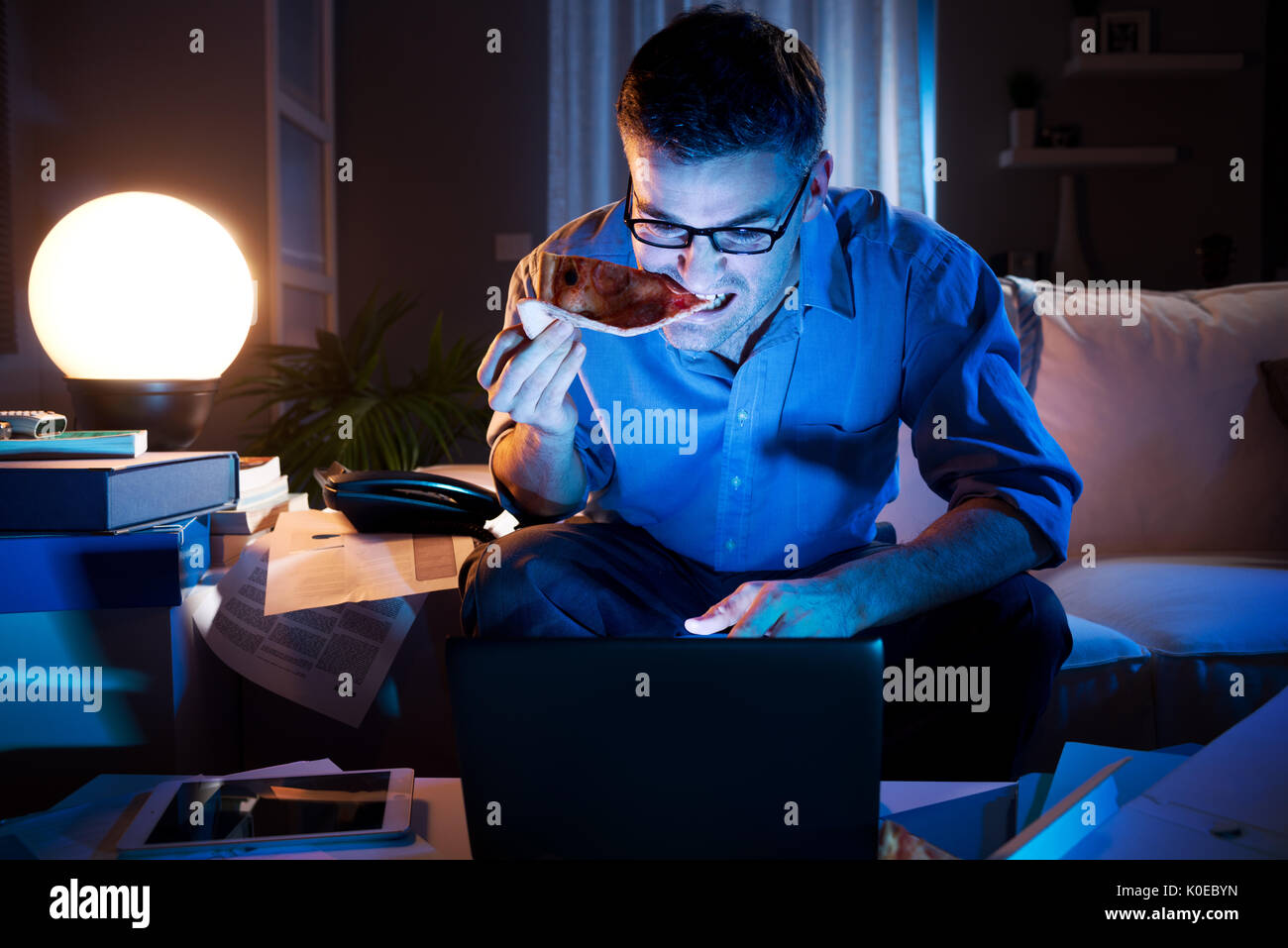 Man eating a slice of pizza and staring at computer screen at home. Stock Photo