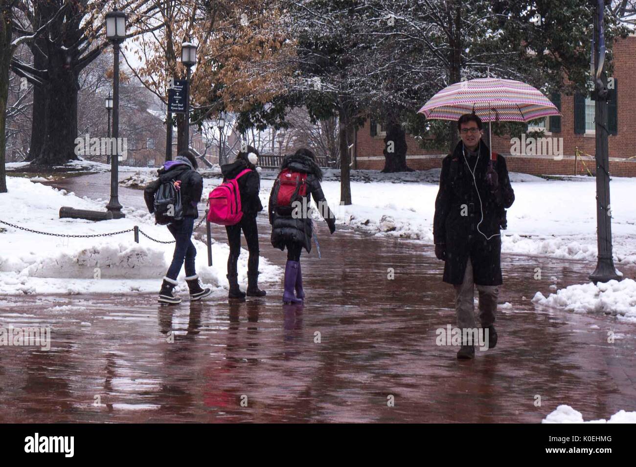 A male graduate student holding an umbrella walks towards the camera as three female undergraduate students in warm clothes and snow boots walk behind him in the opposite direction while it is raining and several inches of snow cover the ground, located between the Freshman Quad and Keyser Quad at Johns Hopkins University, Baltimore, Maryland, December 10, 2013. Courtesy Eric Chen. Stock Photo