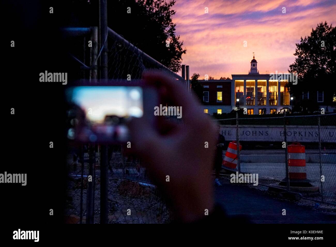 The background of the Johns Hopkins University sign and the Milton S. Eisenhower Library at sunset with a purple and pink clouded sky, with the foreground of a student using their iPhone to take a landscape photo of the sky at Johns Hopkins University, Baltimore, Maryland, October 7, 2013. Courtesy Eric Chen. Stock Photo