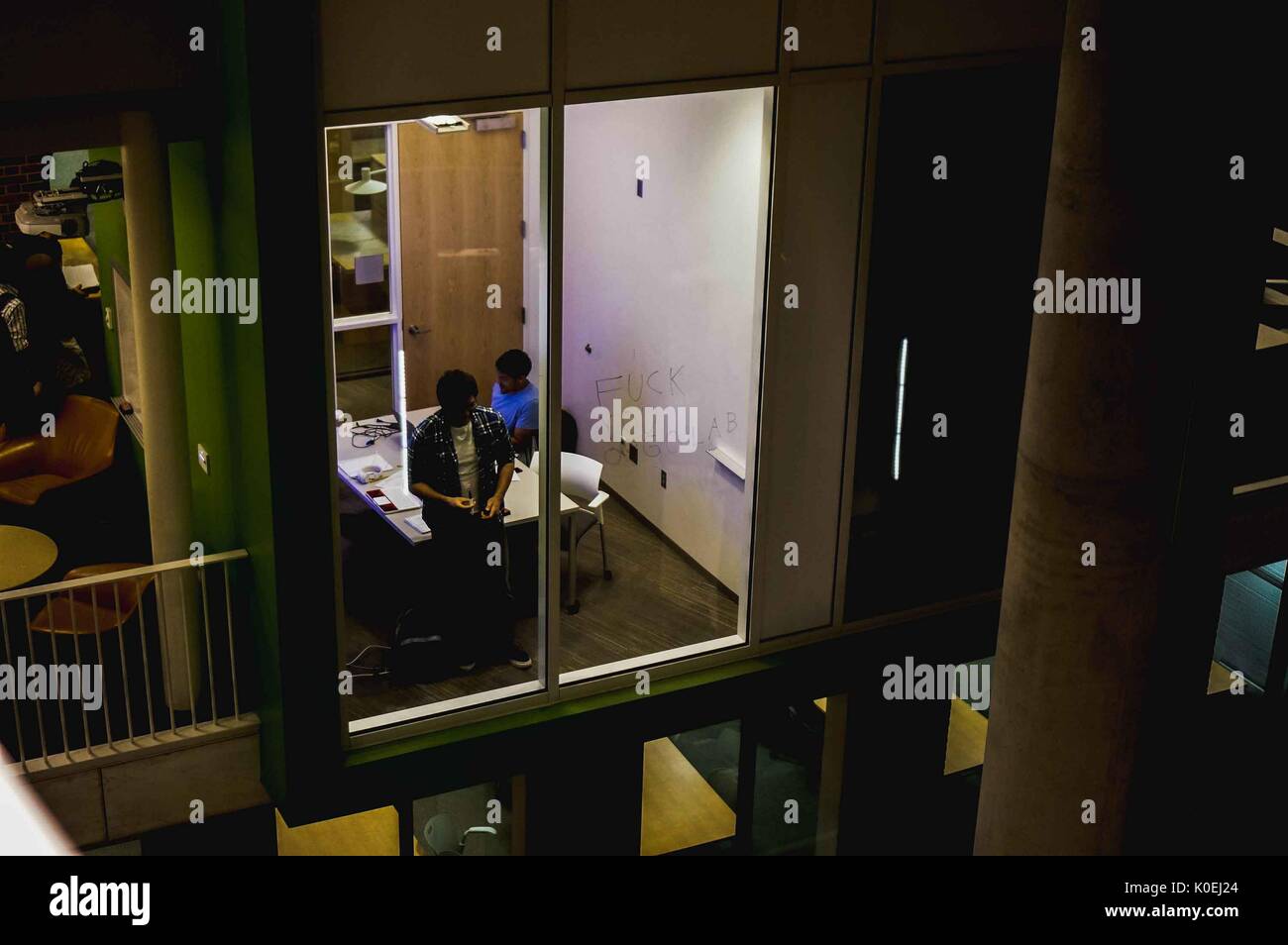 A shadowy shot of two students studying for Organic Chemistry in a cube study room in the Brody Learning Commons at Johns Hopkins University, working out problems on the walls made of white-board and glass windows, an expletive written on the glass; outside of the room, other students work out problems on white-boards and chairs; tables of floors below can be seen amid the architecture of the building's atrium; Baltimore, Maryland, 2014. Courtesy Eric Chen. Stock Photo