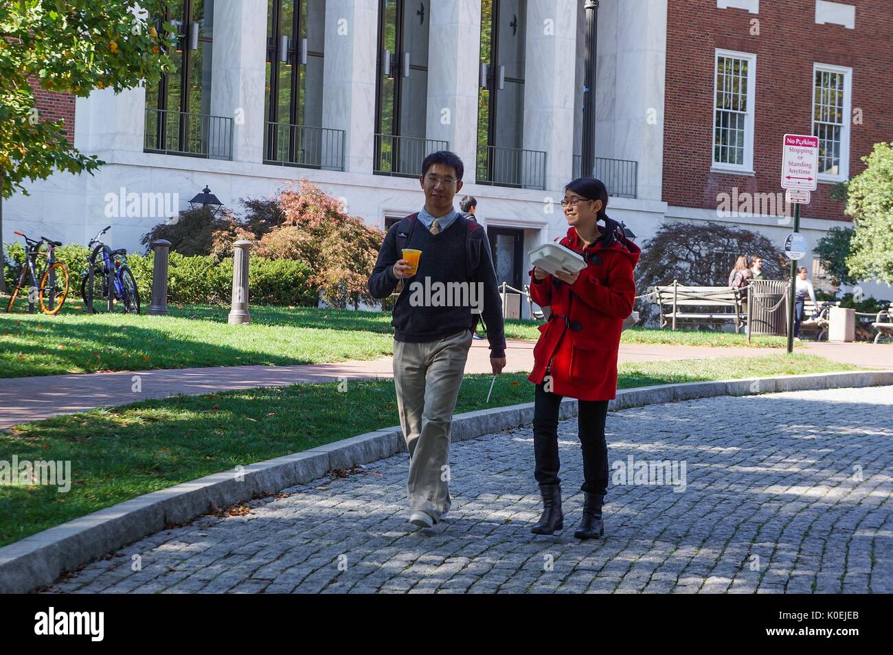 One male and one female college student holding drink and food take a stroll down the cobble stone driveway in front of the Milton S Eisenhower Library at Johns Hopkins University, smiling on a sunny day; bicycles and other students can be seen behind them on the grass in front of the library's entrance; Baltimore, Maryland, March, 2014. Courtesy Eric Chen. Stock Photo