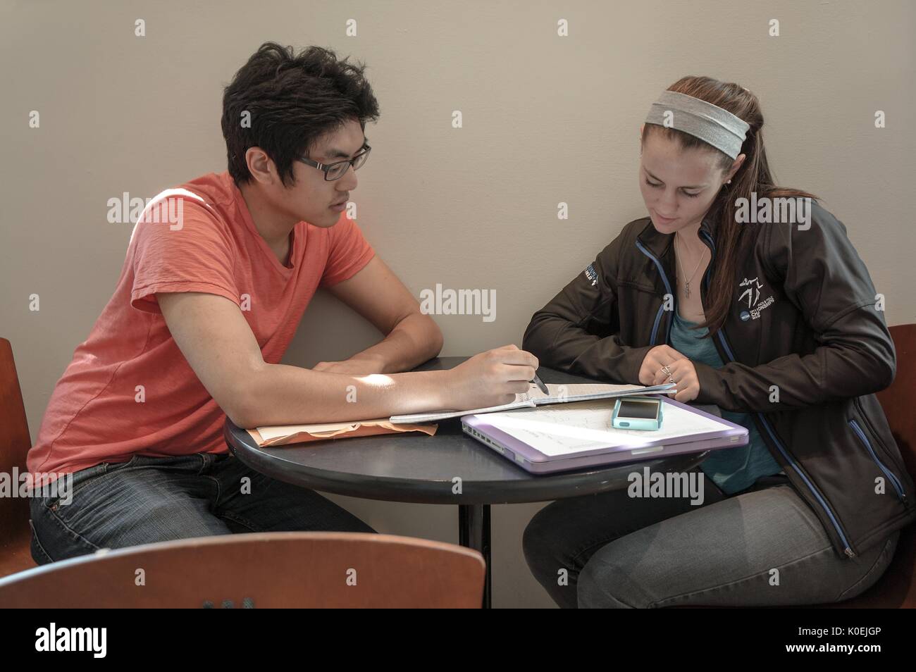 A male student points to notes as a female student listens to his explanation, seated at a small table in the library, a laptop and cellular phone buried under the notebooks; Baltimore, Maryland, March, 2014. Courtesy Eric Chen. Stock Photo