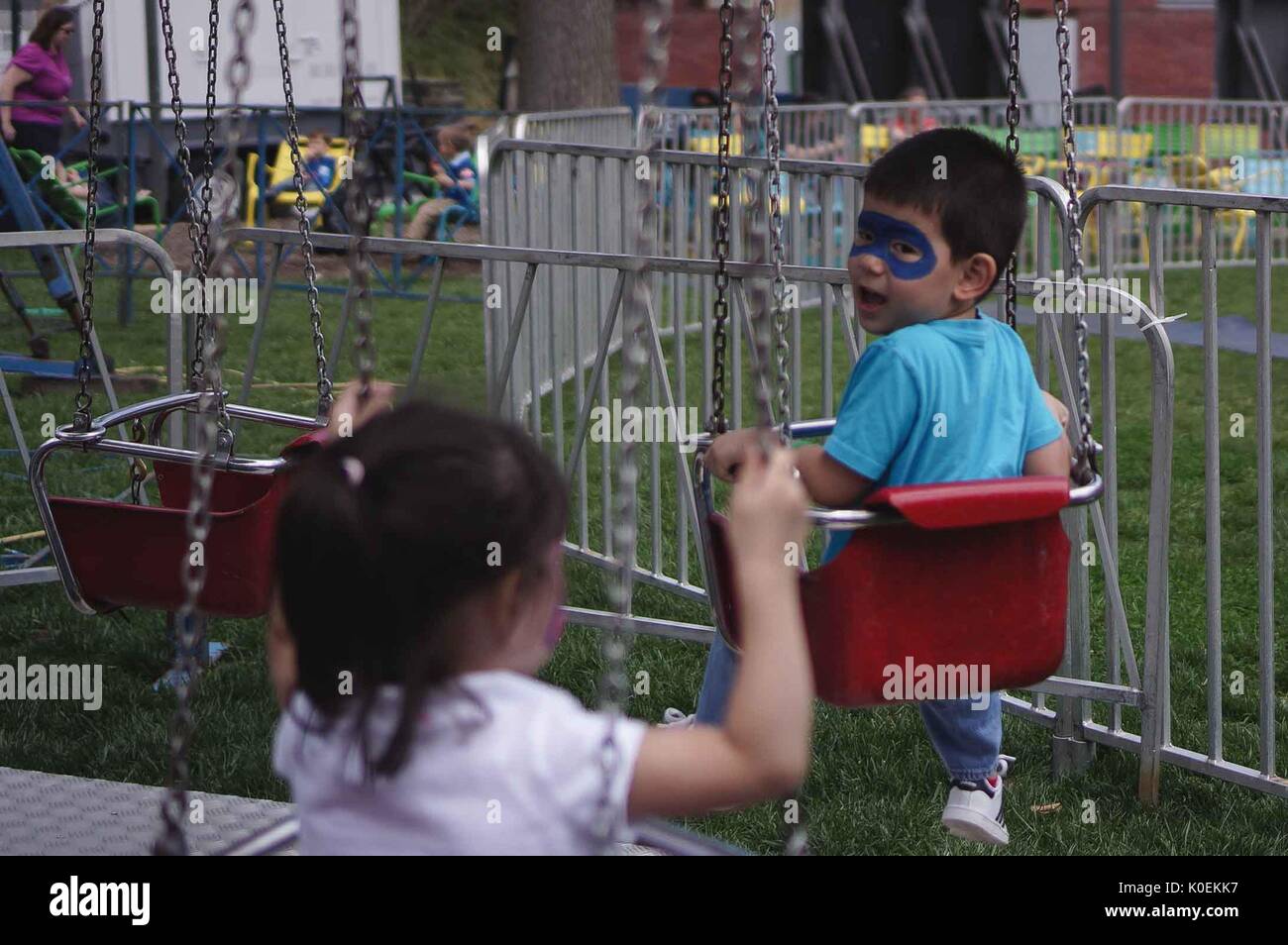 A toddler with face-paint on a small swing ride glances back at his sister in a swing behind him at Spring Fair, a student-run Spring carnival, at Johns Hopkins University, Baltimore, Maryland. April, 2015. Courtesy Eric Chen. Stock Photo