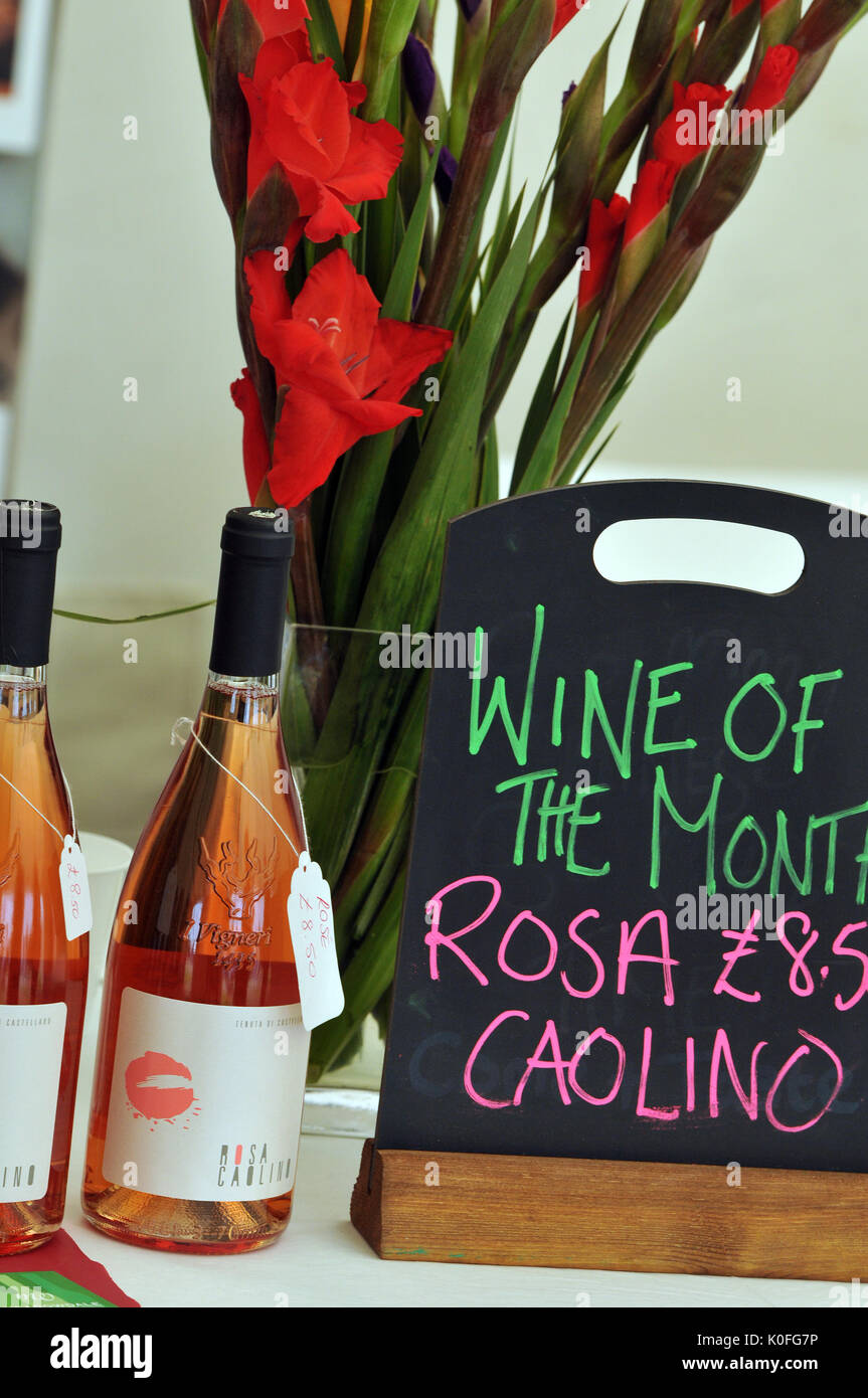 Two bottles of locally produced wine at a farmers market with a sign stating that this is the wine of the month next to some red flowers in a vase. Stock Photo