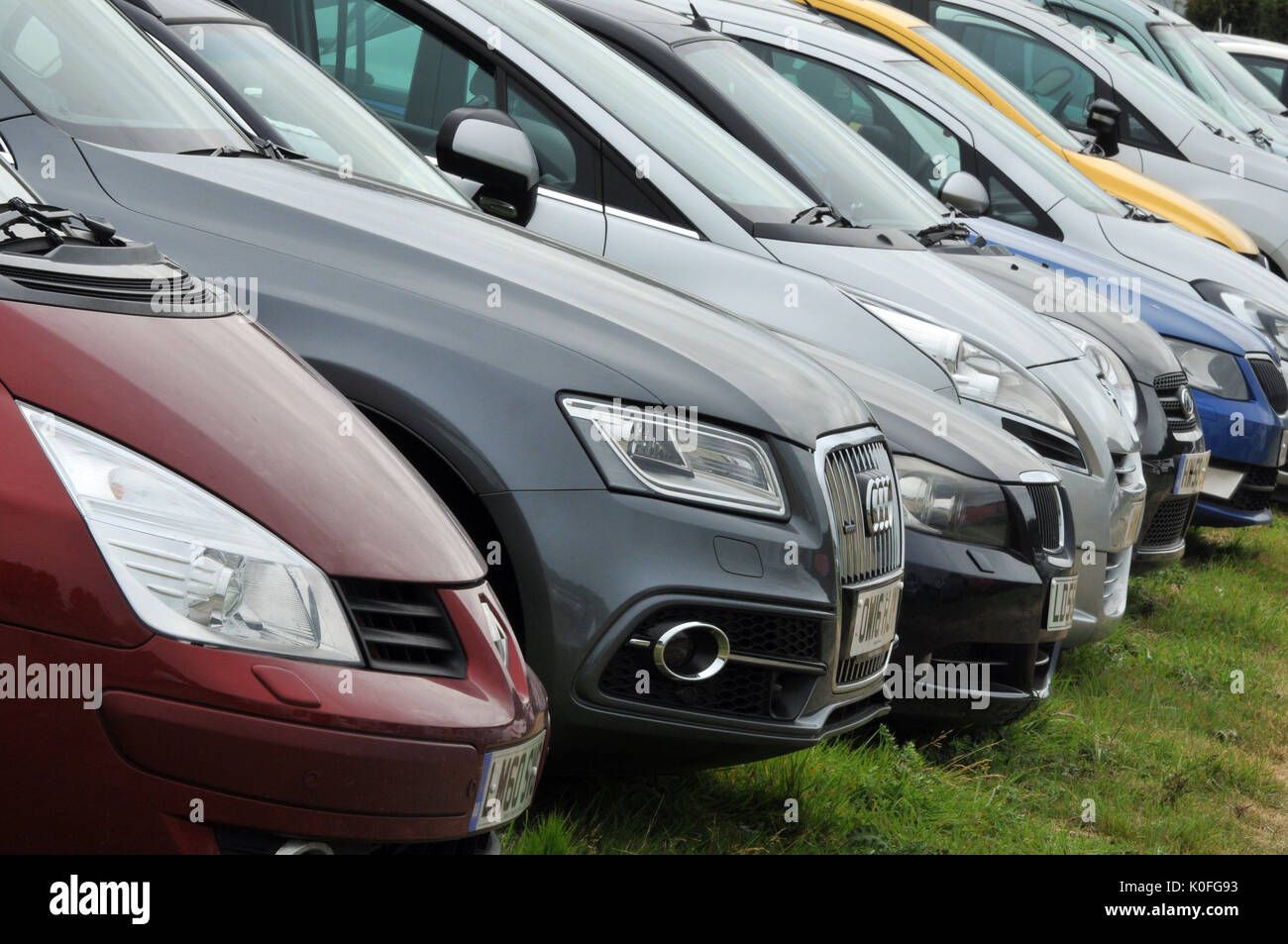 a row or line of modern cars parked in a row in a field or carpark different colours headlights and bumpers on the grass temporary car parking Stock Photo