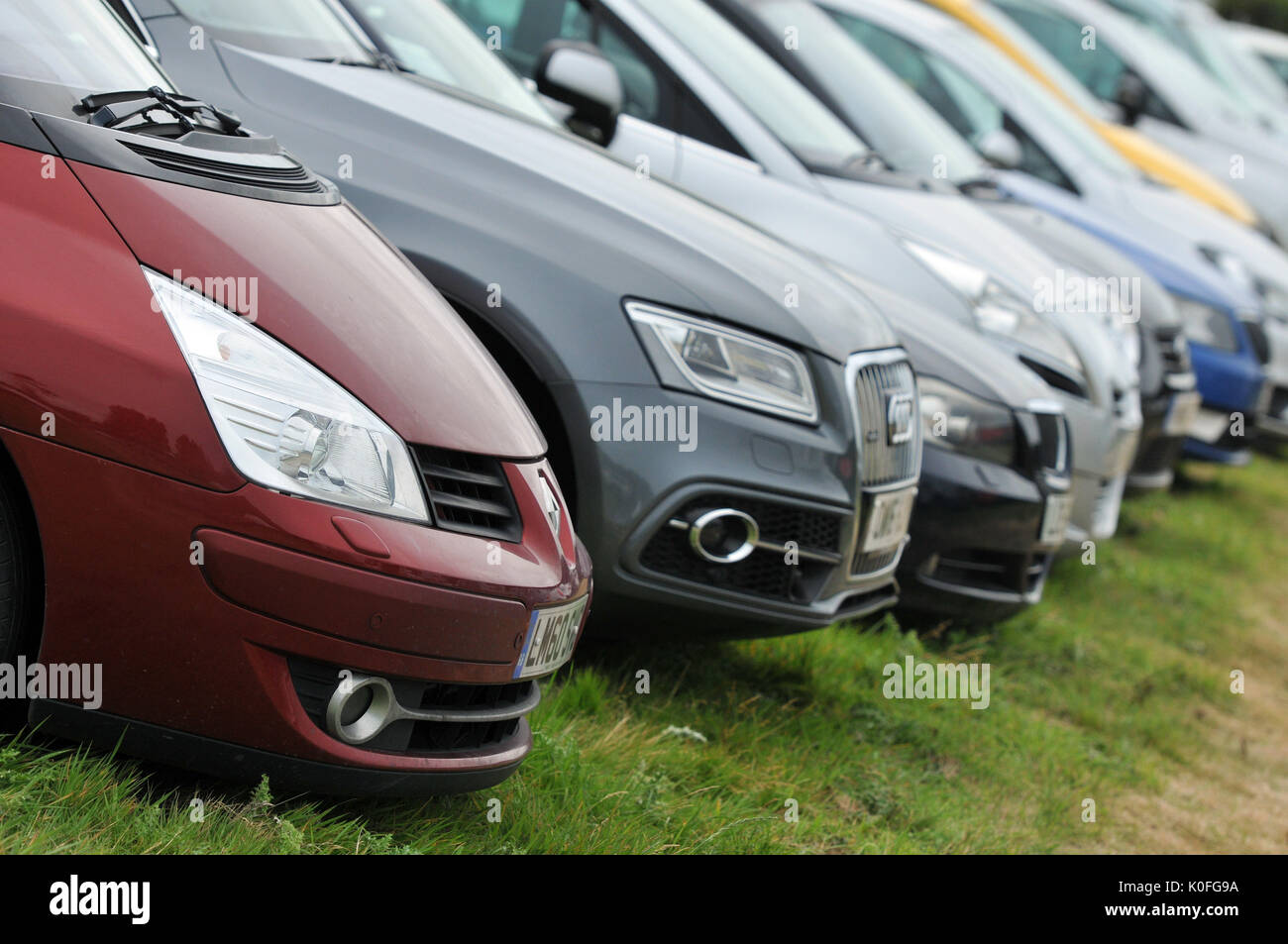 a row or line of modern cars parked in a row in a field or carpark different colours headlights and bumpers on the grass temporary car parking Stock Photo