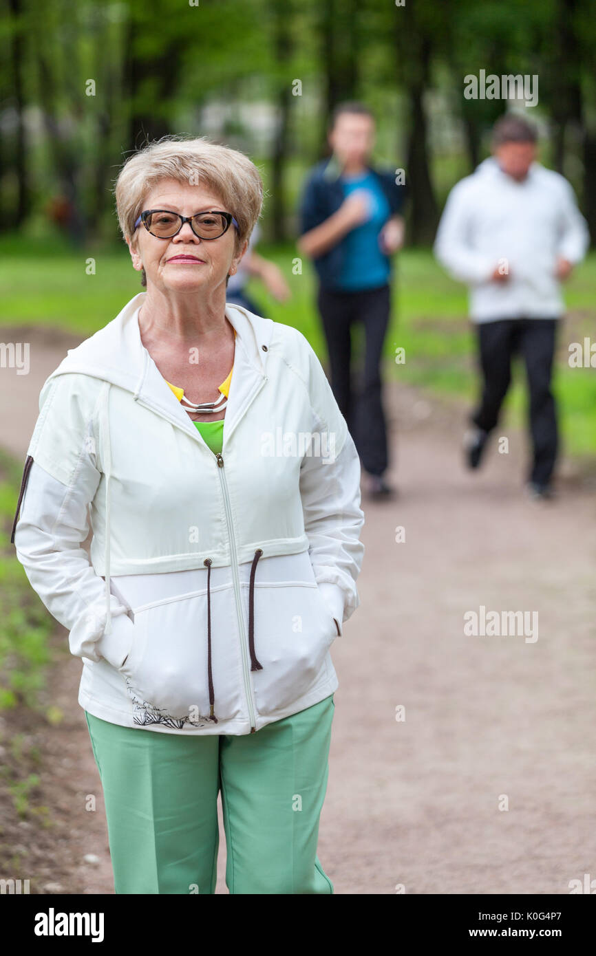 European elderly woman in a sports suit standing in the park on background of running people Stock Photo