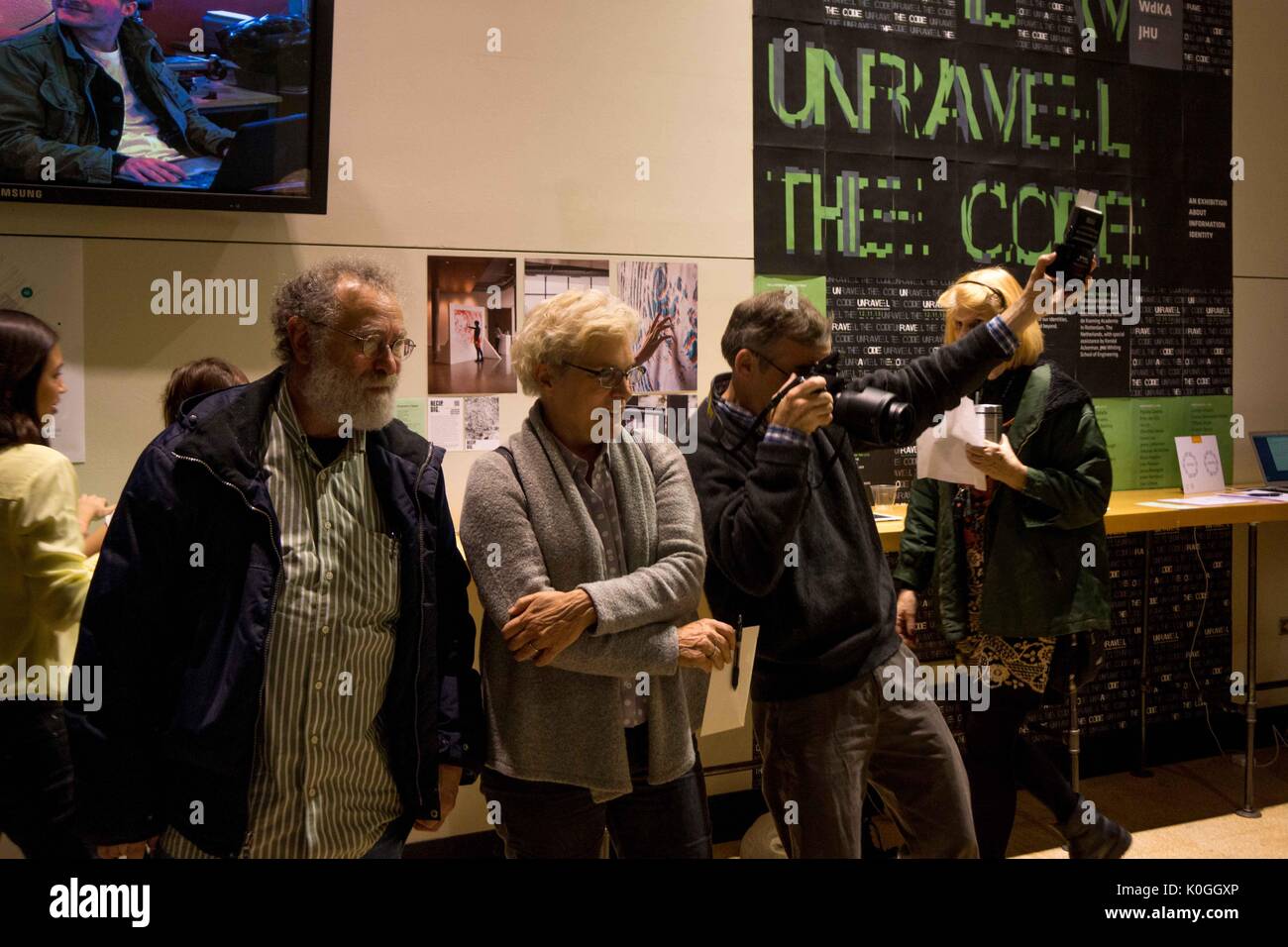 Two attendees look on at artwork not pictured while one man takes a photo of the artwork at the Unravel the Code Opening at The Johns Hopkins University Sheridan Libraries, 2016. Courtesy Eric Chen. Stock Photo