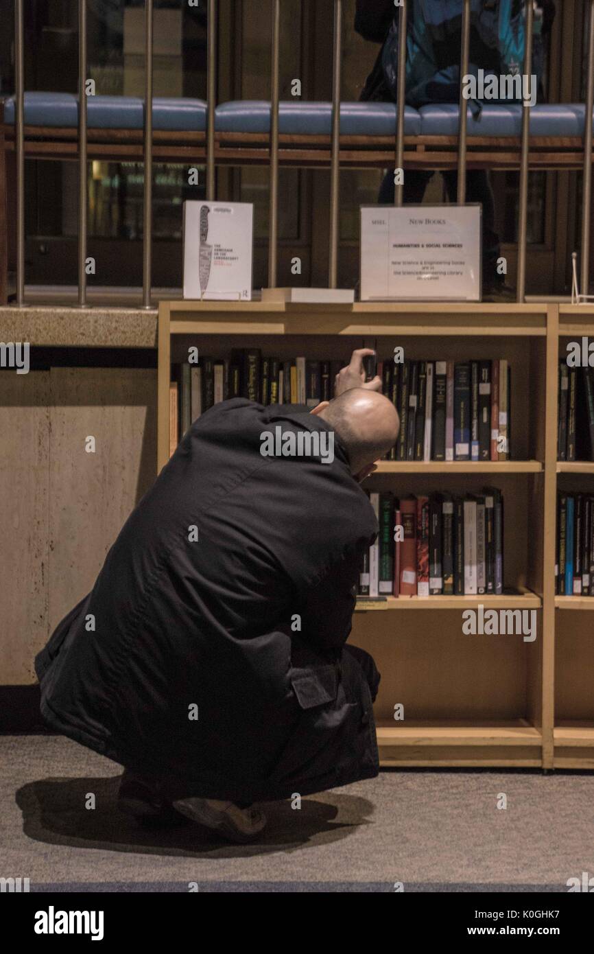 A man squats and tilts his head, reading the labels on the bindings of books on a shelf and drawing one out, in the Milton S. Eisenhower Library on the Homewood campus of the Johns Hopkins University in Baltimore, Maryland, 2014. Courtesy Eric Chen. Stock Photo