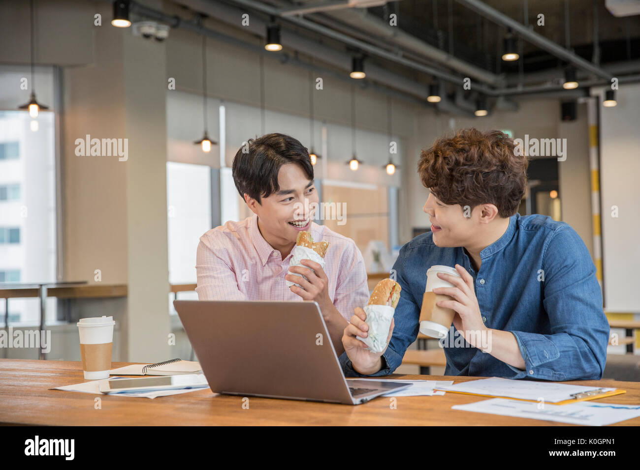 Portrait of two smiling businessmen with sandwiches having a talk sharing a notebook computer at cafeteria Stock Photo
