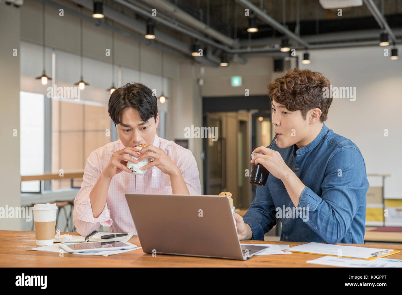 Two businessmen having coffee and sandwiches sharing a notebook computer at cafeteria Stock Photo