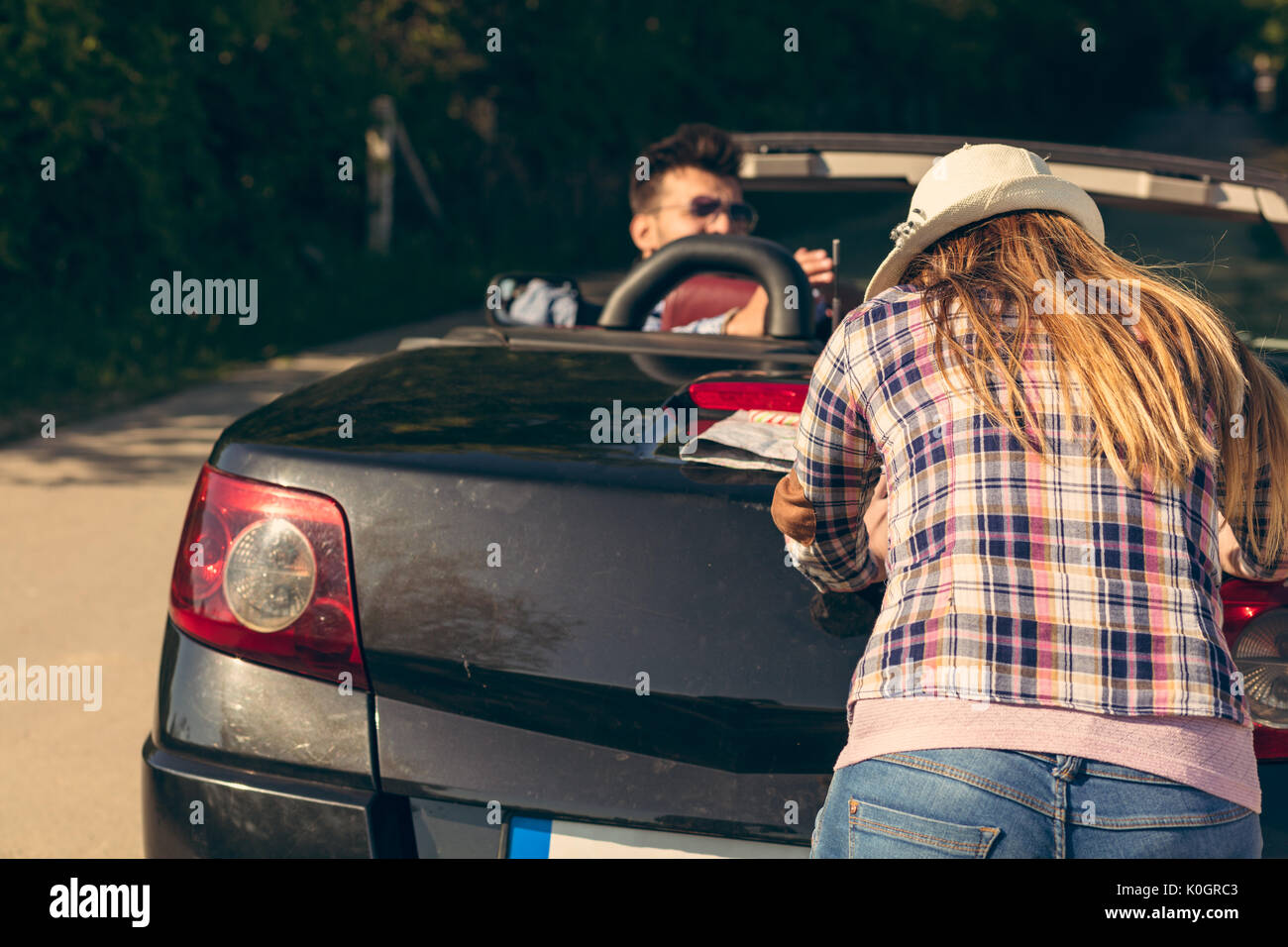 leisure, road trip, travel and people concept - happy friends pushing broken cabriolet car along country road Stock Photo