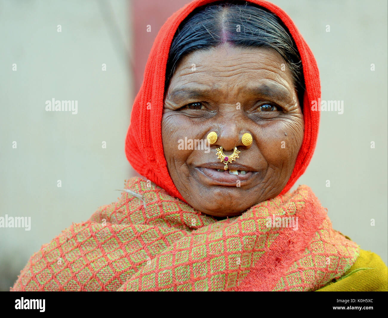 Elderly Indian Adivasi woman with two golden nose studs and gold-and-gemstone nose jewellery wears a red headscarf and smiles for the camera. Stock Photo