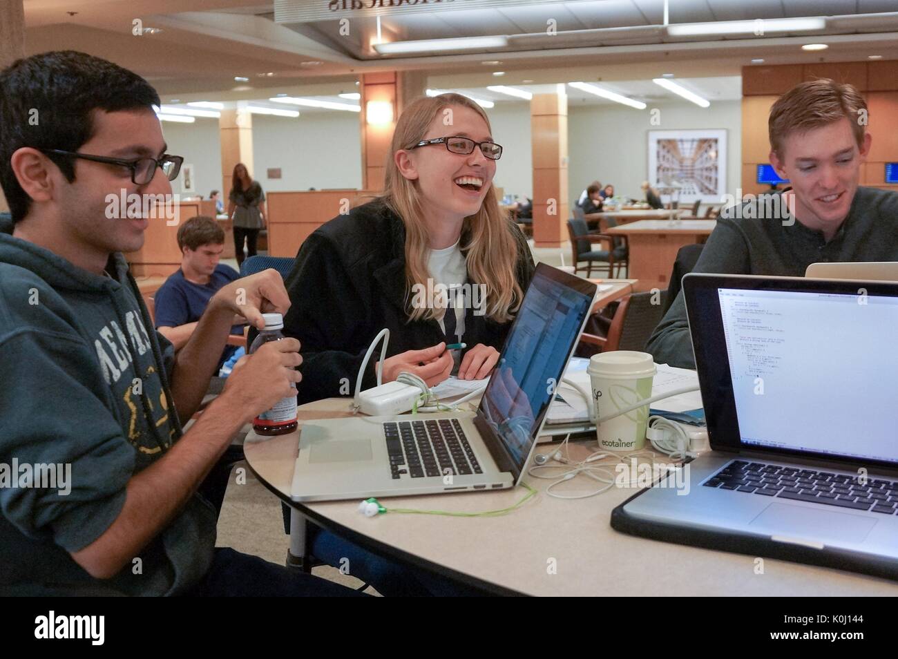 Three students, two males on either side of a female, seated on a table in the second level, M Level, of the Milton S. Eisenhower Library, surrounded by study materials and laptops, other students depicted in the background, all with smiling facial expressions, 2016. Courtesy Eric Chen. Stock Photo
