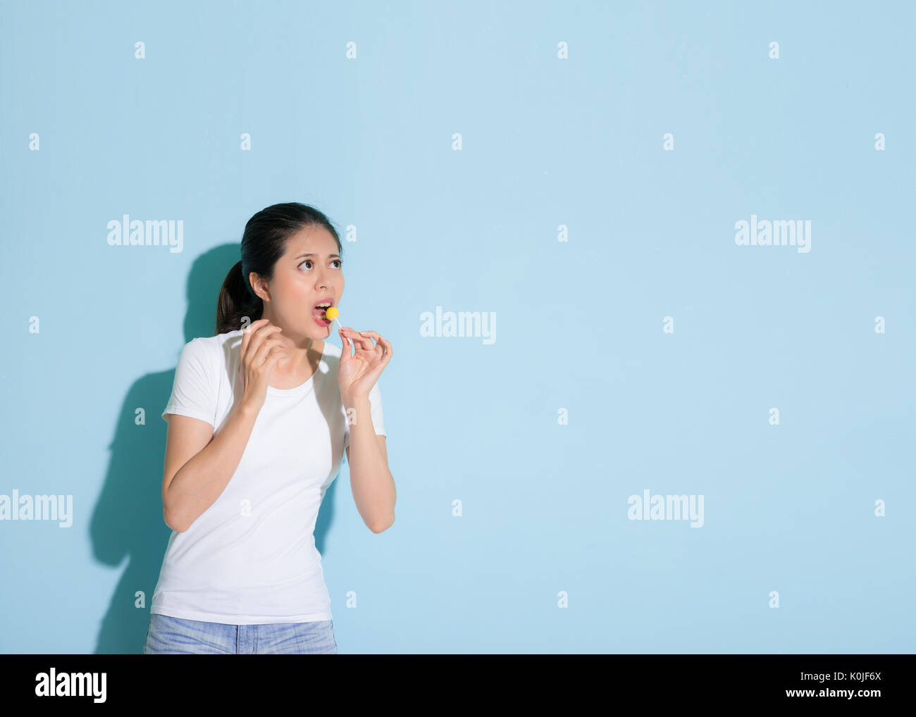 cute beautiful female model eating lollipop and looking empty area worry about tooth decay problem performance in blue wall background. Stock Photo