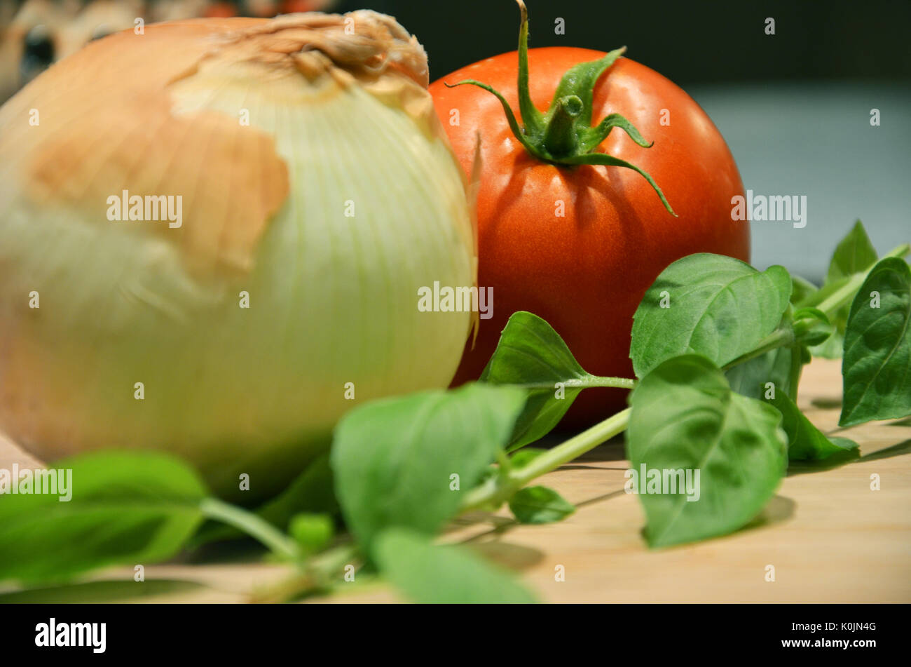 A yellow onion, tomato, and stem of basil on a cutting board in a kitchen as ingredients ready to be cut to make a pasta sauce. Stock Photo