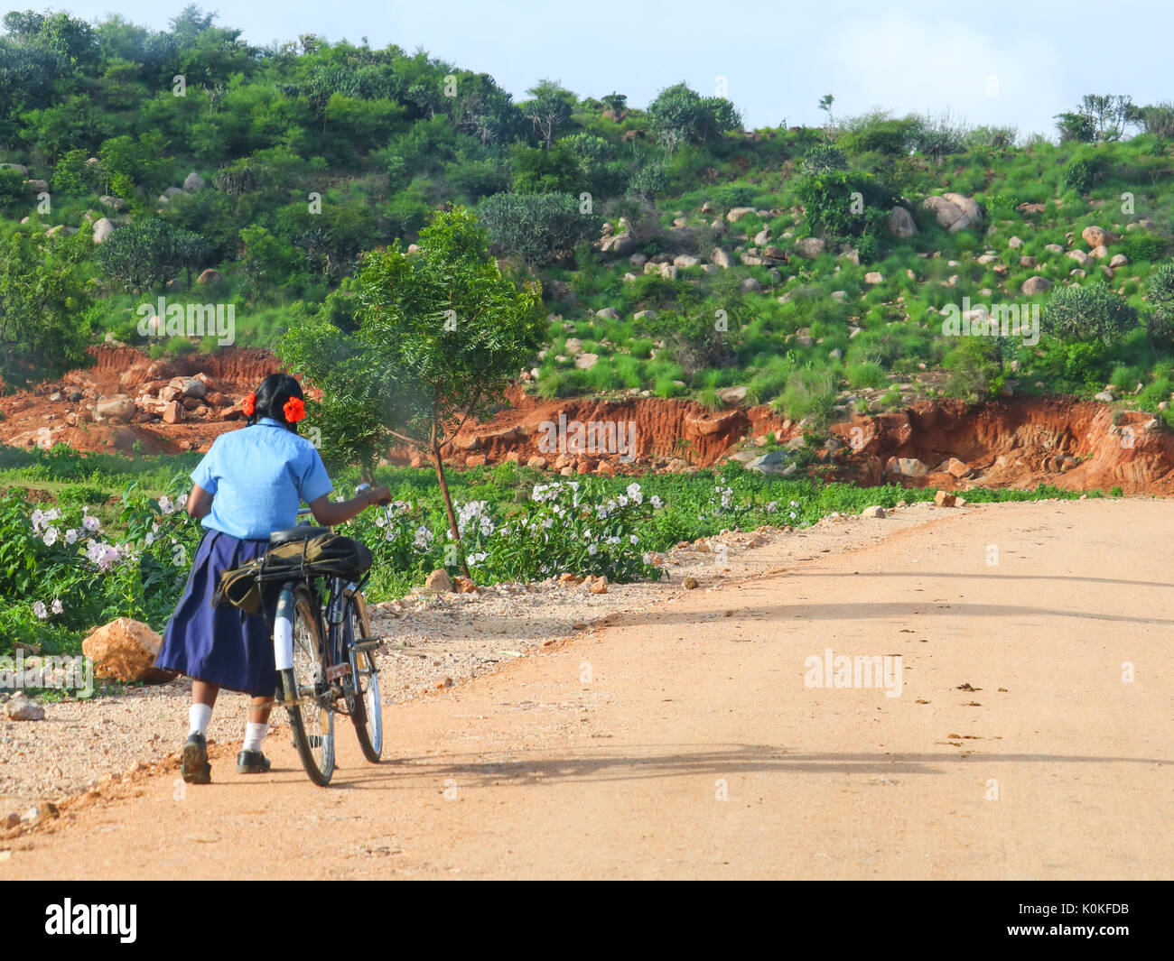 A girl child going to school on a sunny day with her bicycle. A school bag of the child can also be seen in the picture. Stock Photo
