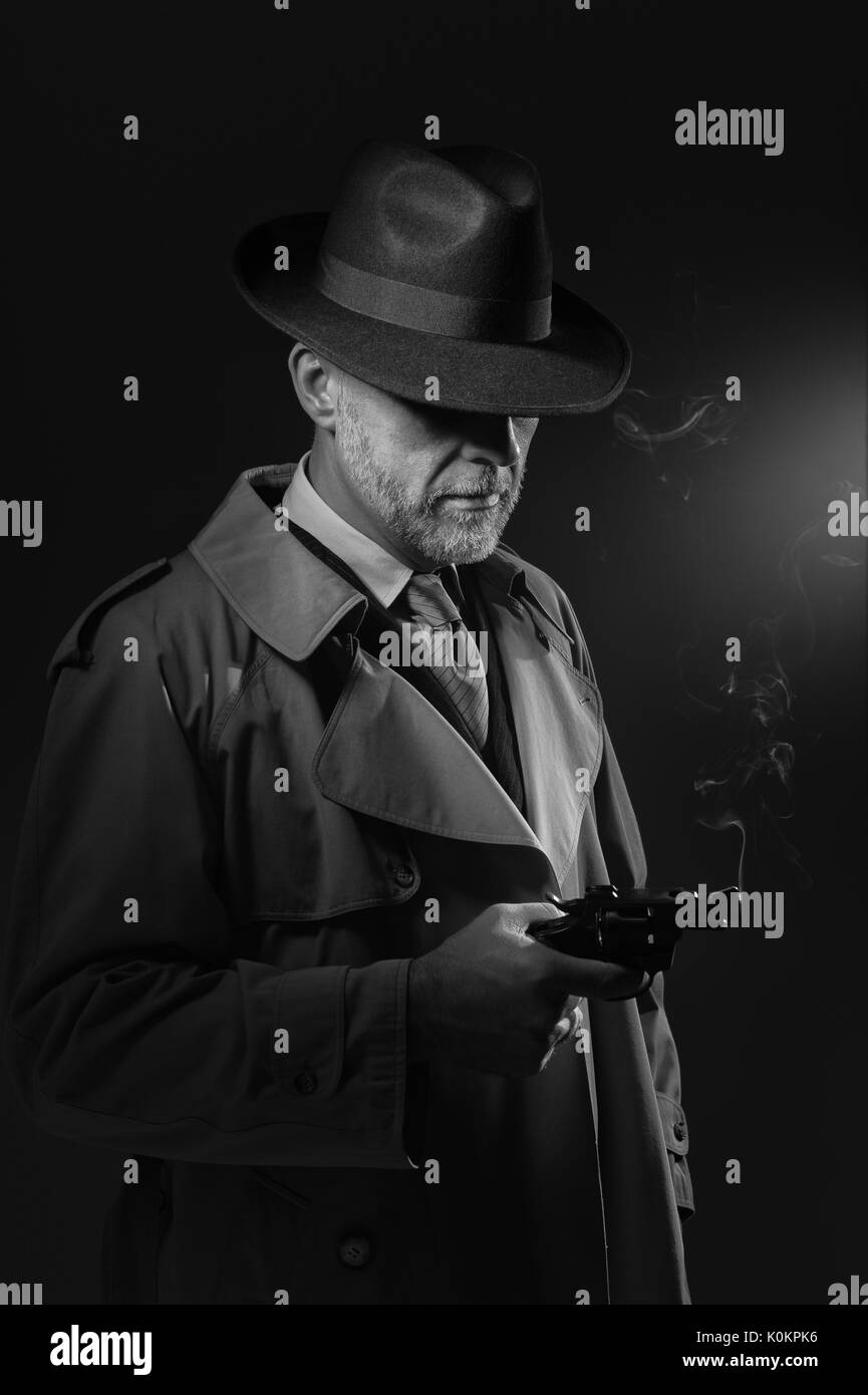 Detective holding a gun, he is wearing a fedora hat and a trench coat, 1950s noir film character Stock Photo