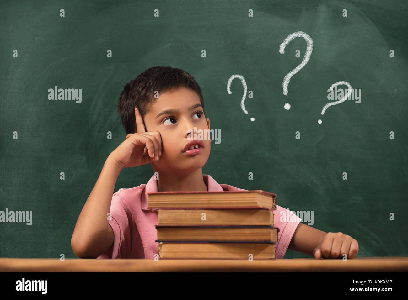 Boy with doubts and thoughts in class. Portrait of male child thinking against question marks on blackboard Stock Photo