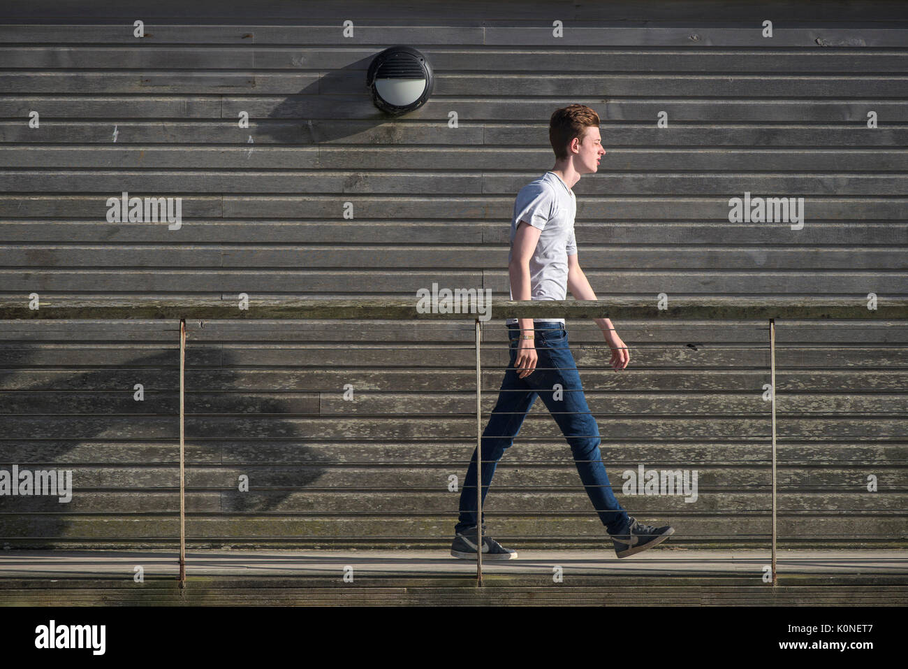 A male teenager walking along a walkway next to a wooden building. Stock Photo